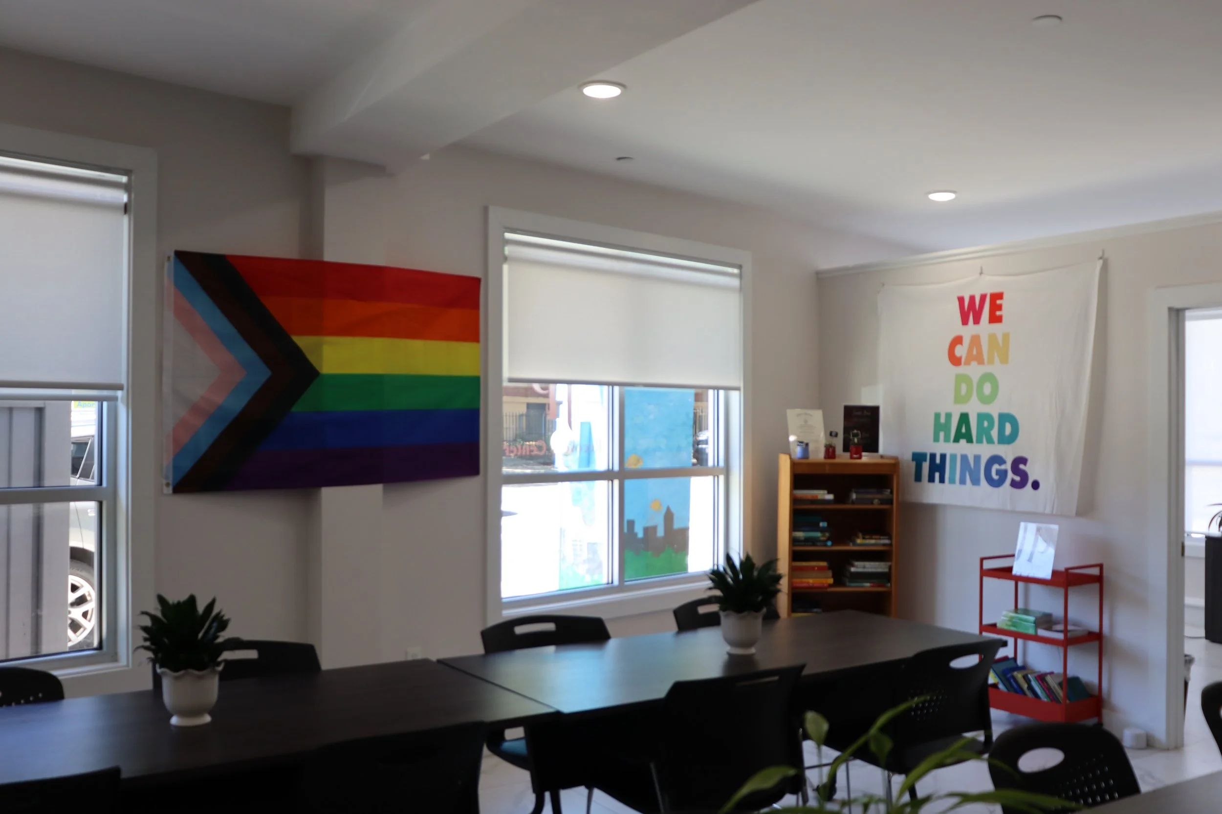 A room with white walls, decorated with an LGBTQ+ pride flag and a banner that reads 'We Can Do Hard Things' in rainbow colors. There are windows, a table with black chairs, and a bookshelf.