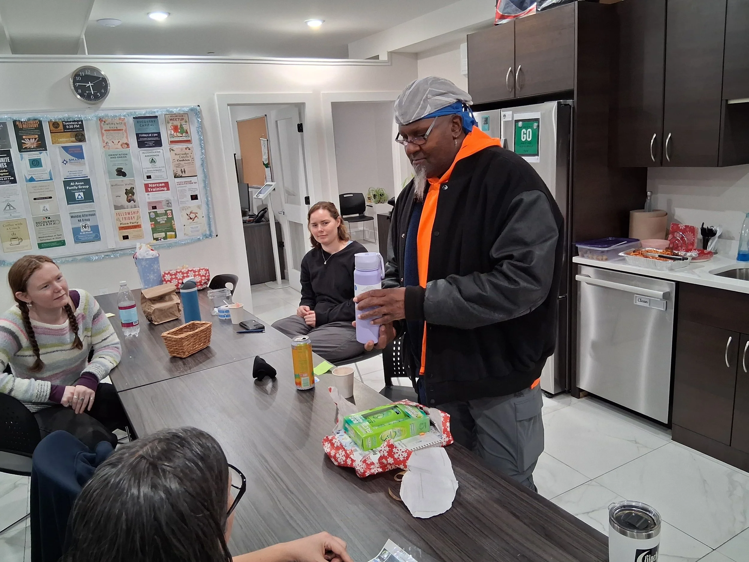 A man standing at a table holding a container, surrounded by young women seated at the table in a kitchen or community room.