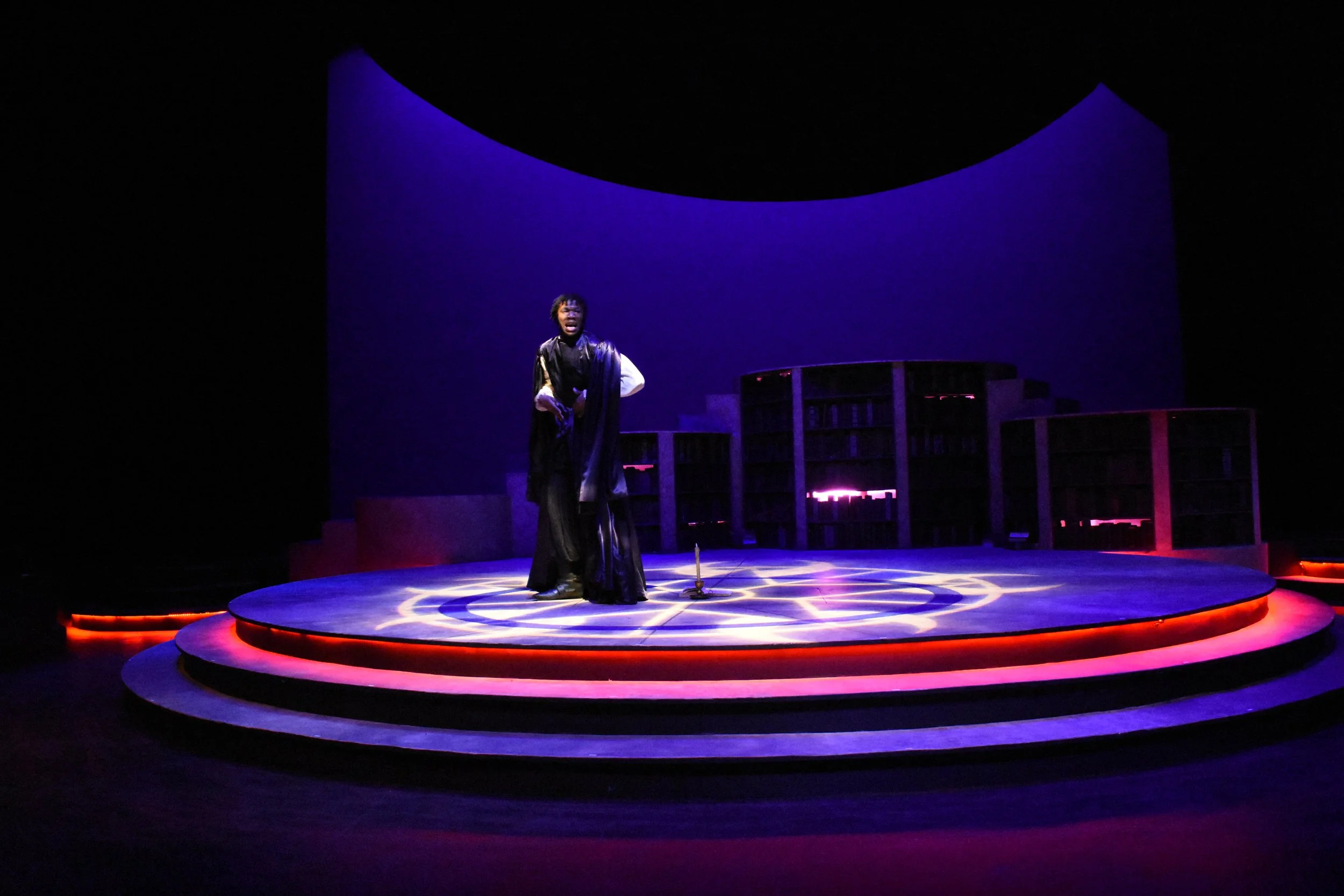 A person standing on a circular stage illuminated with purple and pink lighting in a theatrical setting, with shelves and a curved backdrop behind.