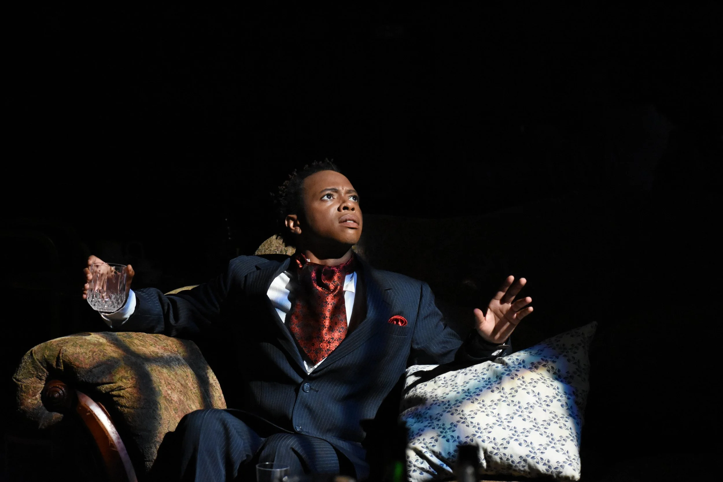 A woman in a dark suit with a red scarf and pocket square sits on a vintage armchair, holding a glass of water, illuminated against a dark background, with a contemplative expression and hand raised.