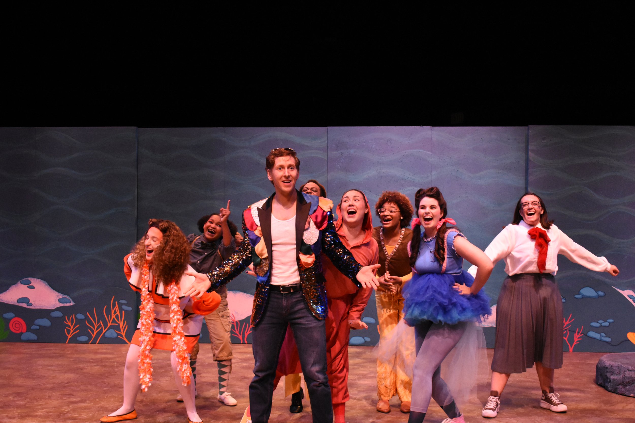 Actors performing on stage in colorful costumes, smiling and laughing, with painted backdrop depicting water and rocks.