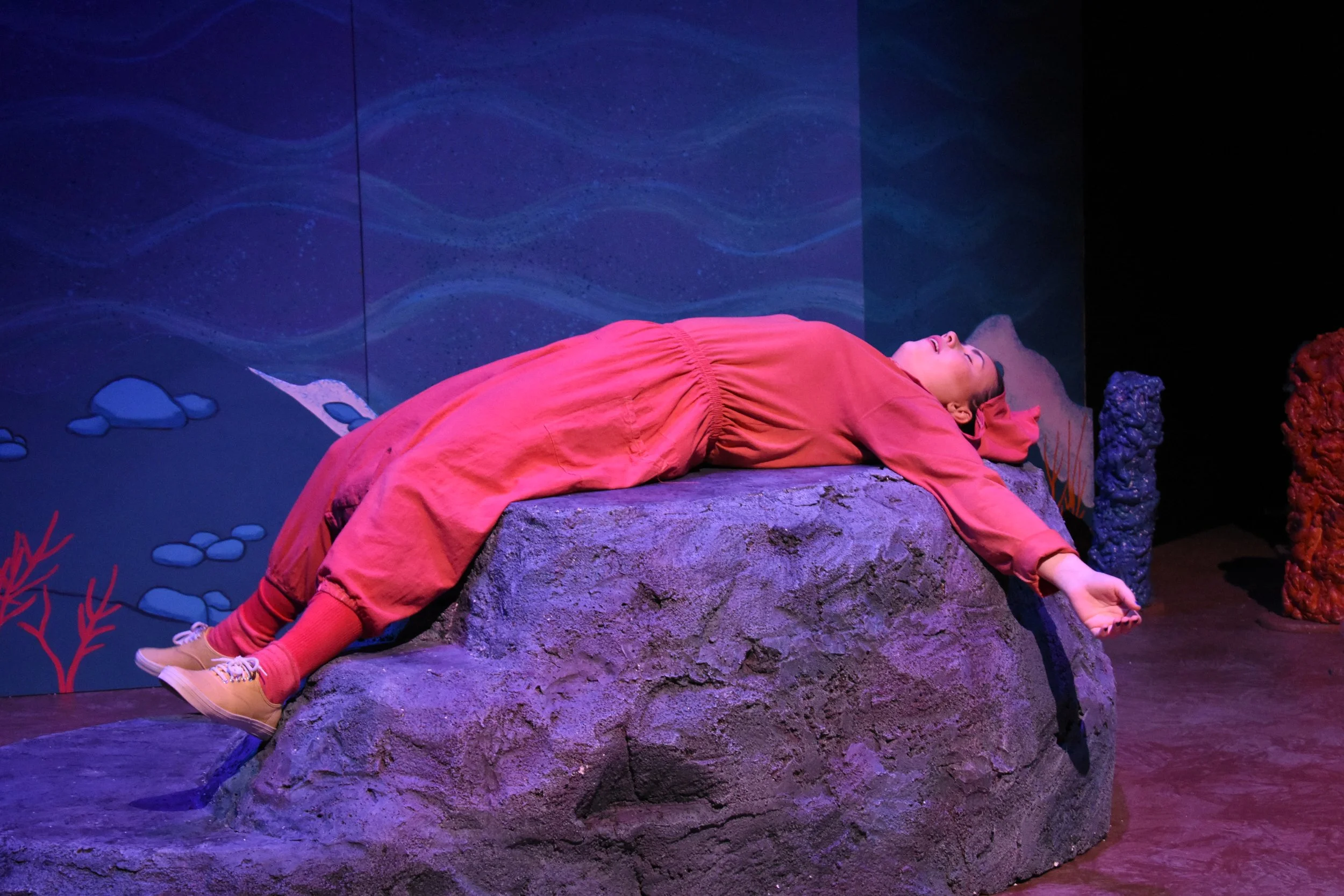 A person lying on a large artificial rock, covered in a pink outfit, with eyes closed, in a colorful underwater-themed setting.
