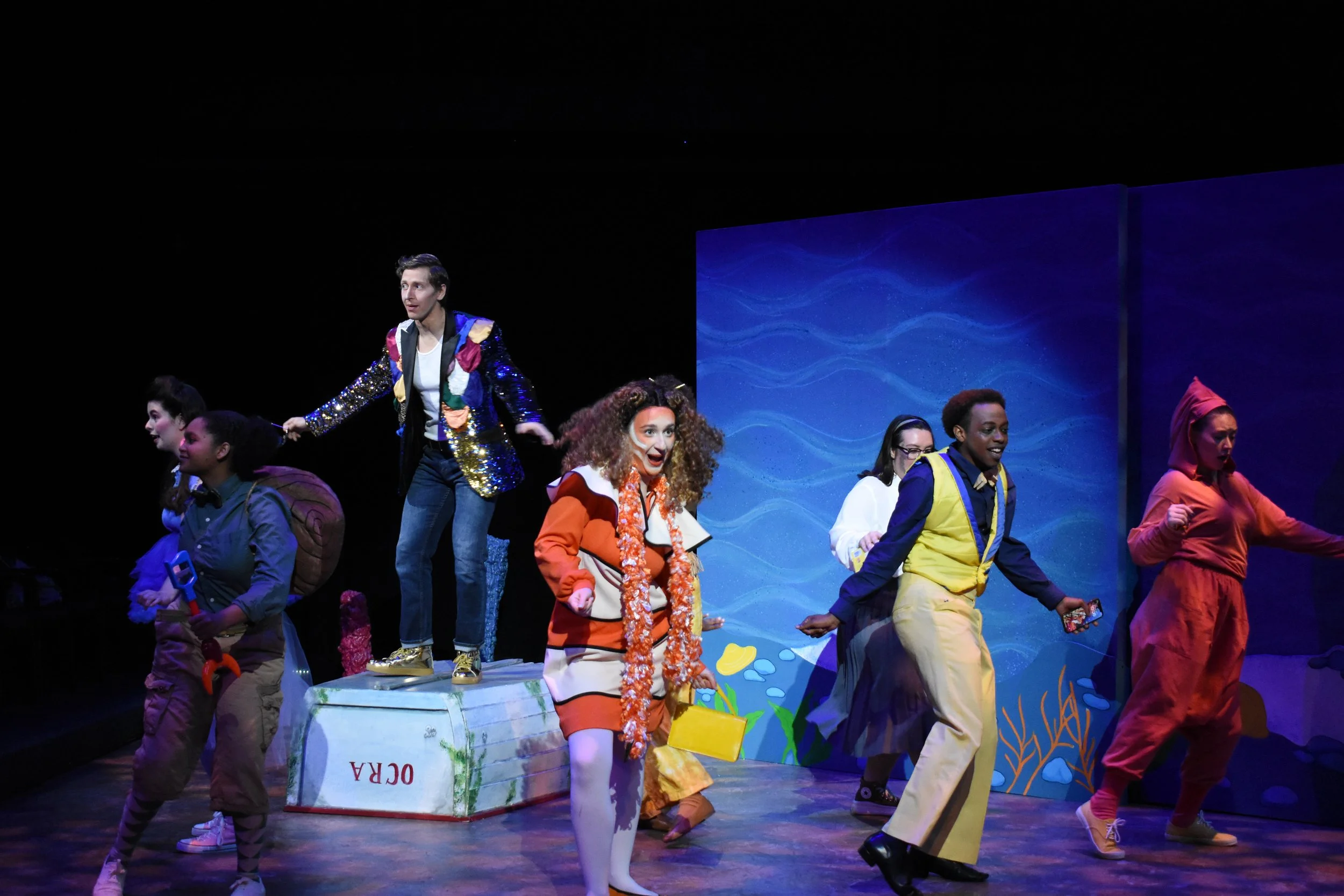 The cast of a theatrical performance on stage, with colorful costumes including a person in a sparkly jacket and others in vibrant outfits, against a blue ocean-themed backdrop.