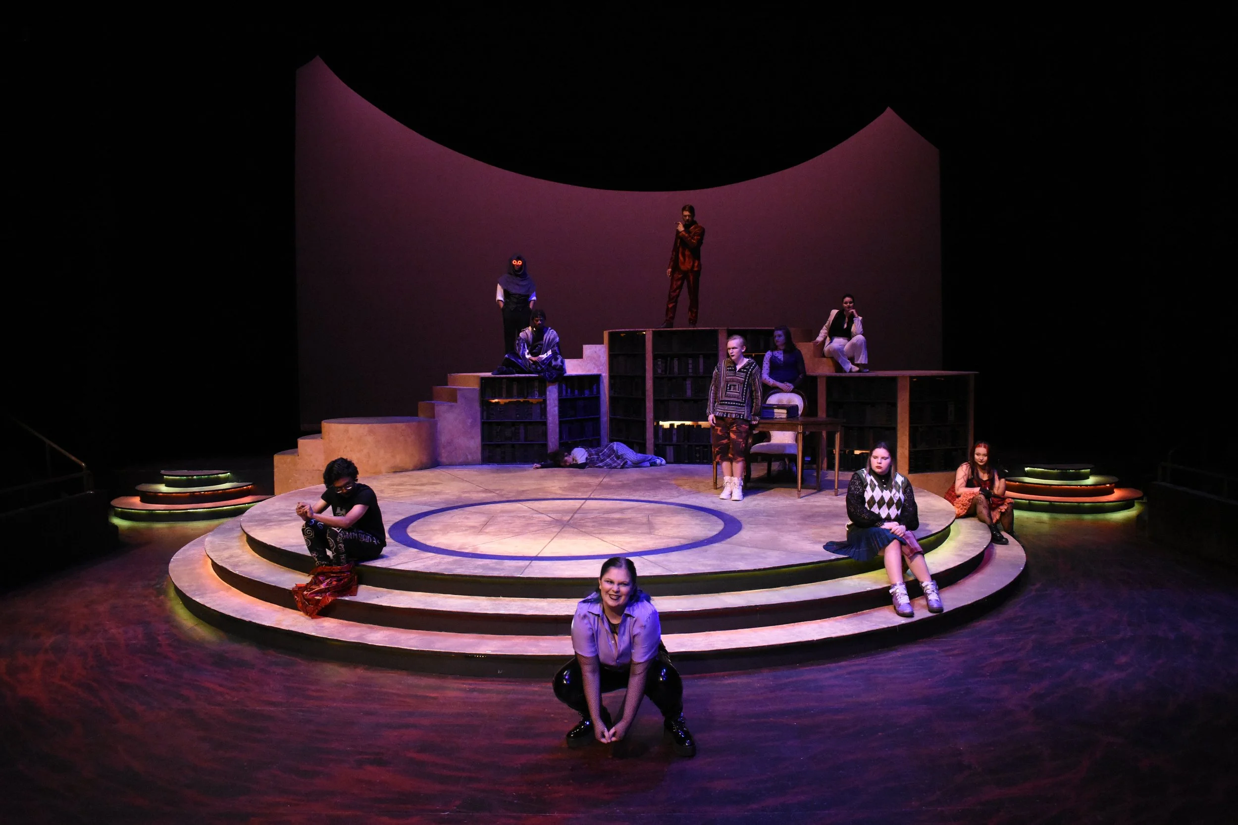 Theater stage with multiple actors, some sitting on steps and others standing, lit with purple and yellow lights