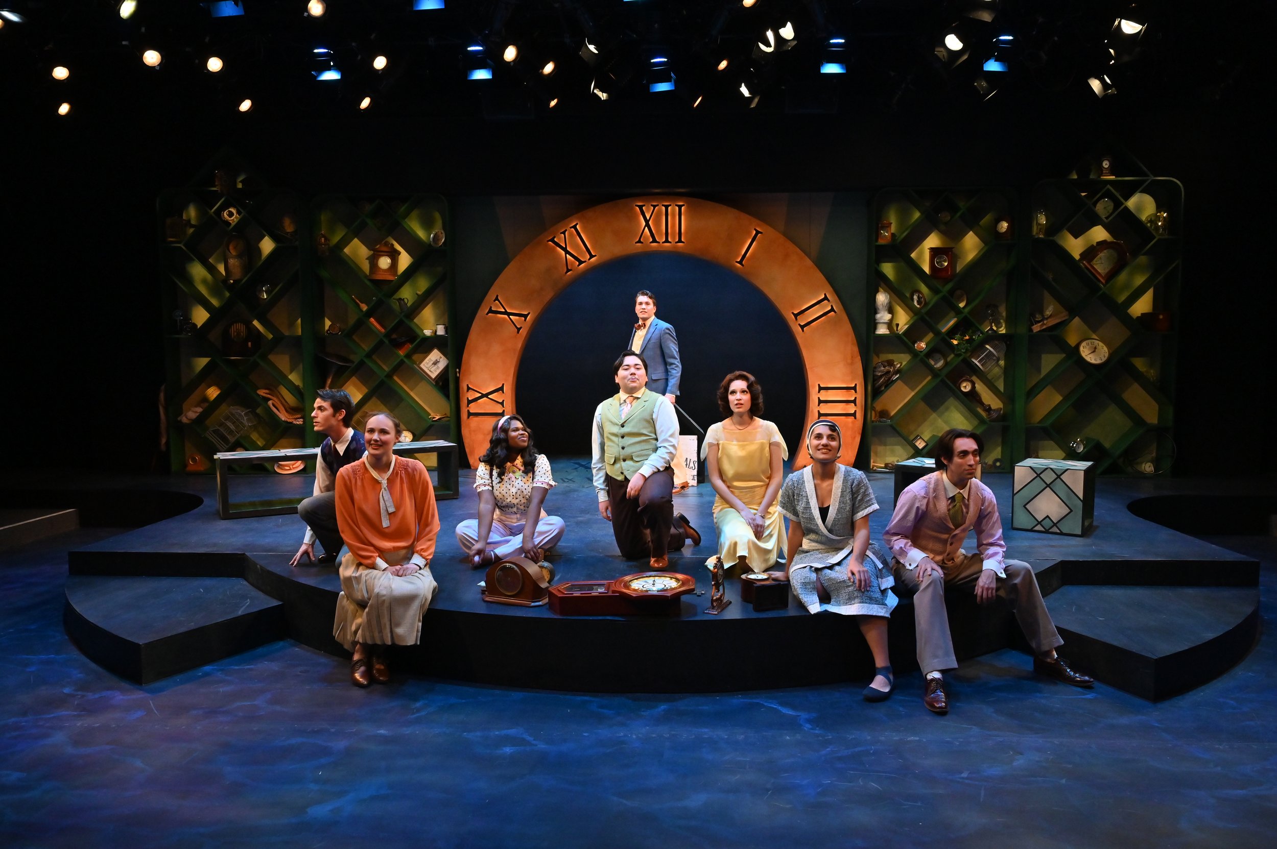 The stage with multiple actors performing a scene in a theater, featuring a large clock face with Roman numerals in the background, and shelves with various objects on either side.