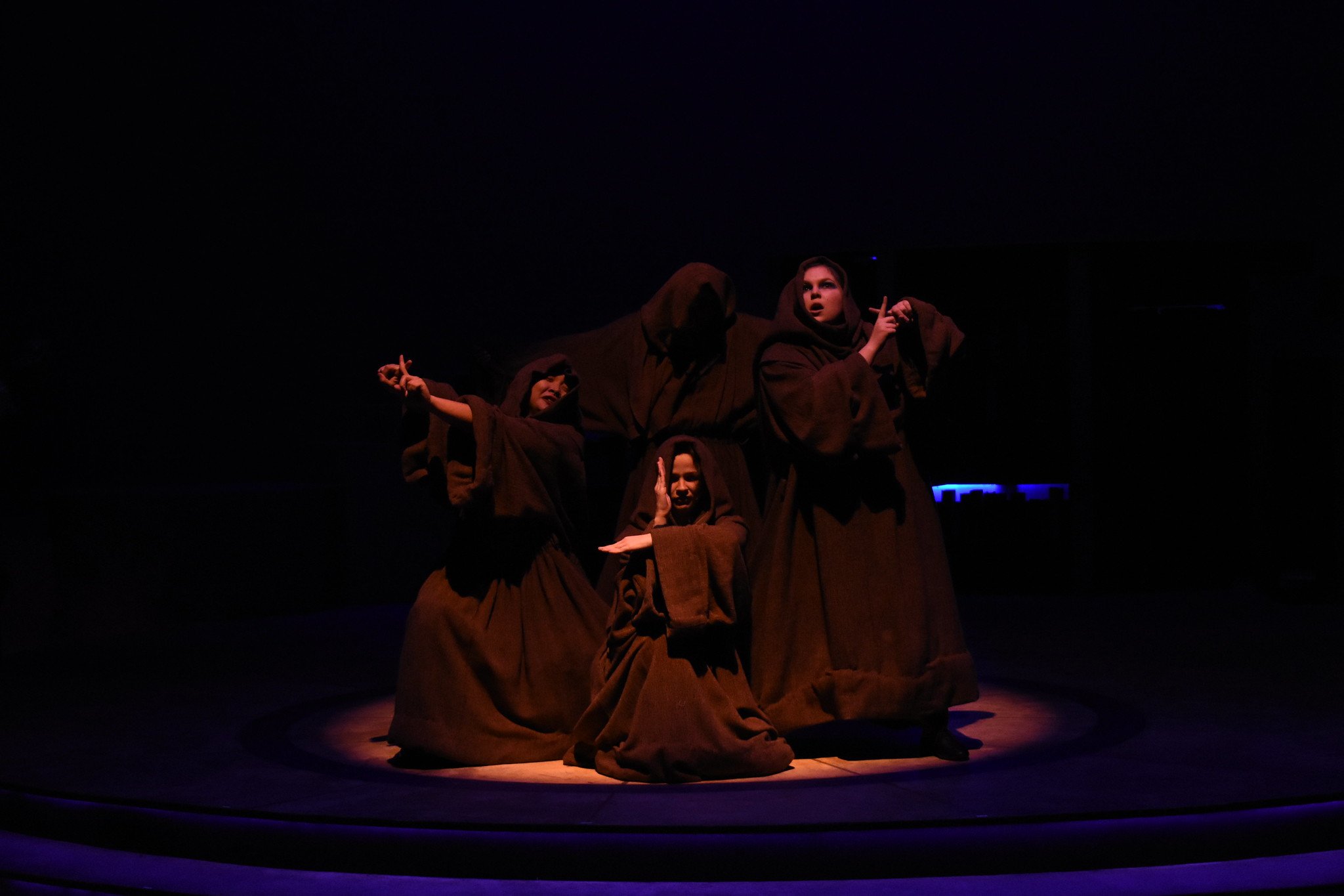 Four performers dressed in brown robes on theater stage, one kneeling, two standing and gesturing, one with a serious expression, in low lighting.