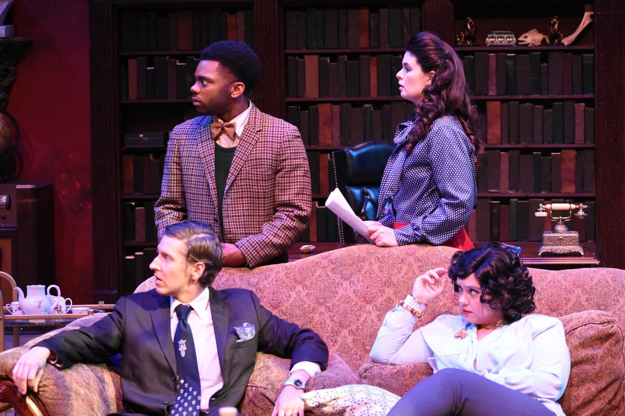 Four people in vintage clothing in a room with a large bookshelf; two are seated on a sofa and two are standing behind it, engaged in conversation.