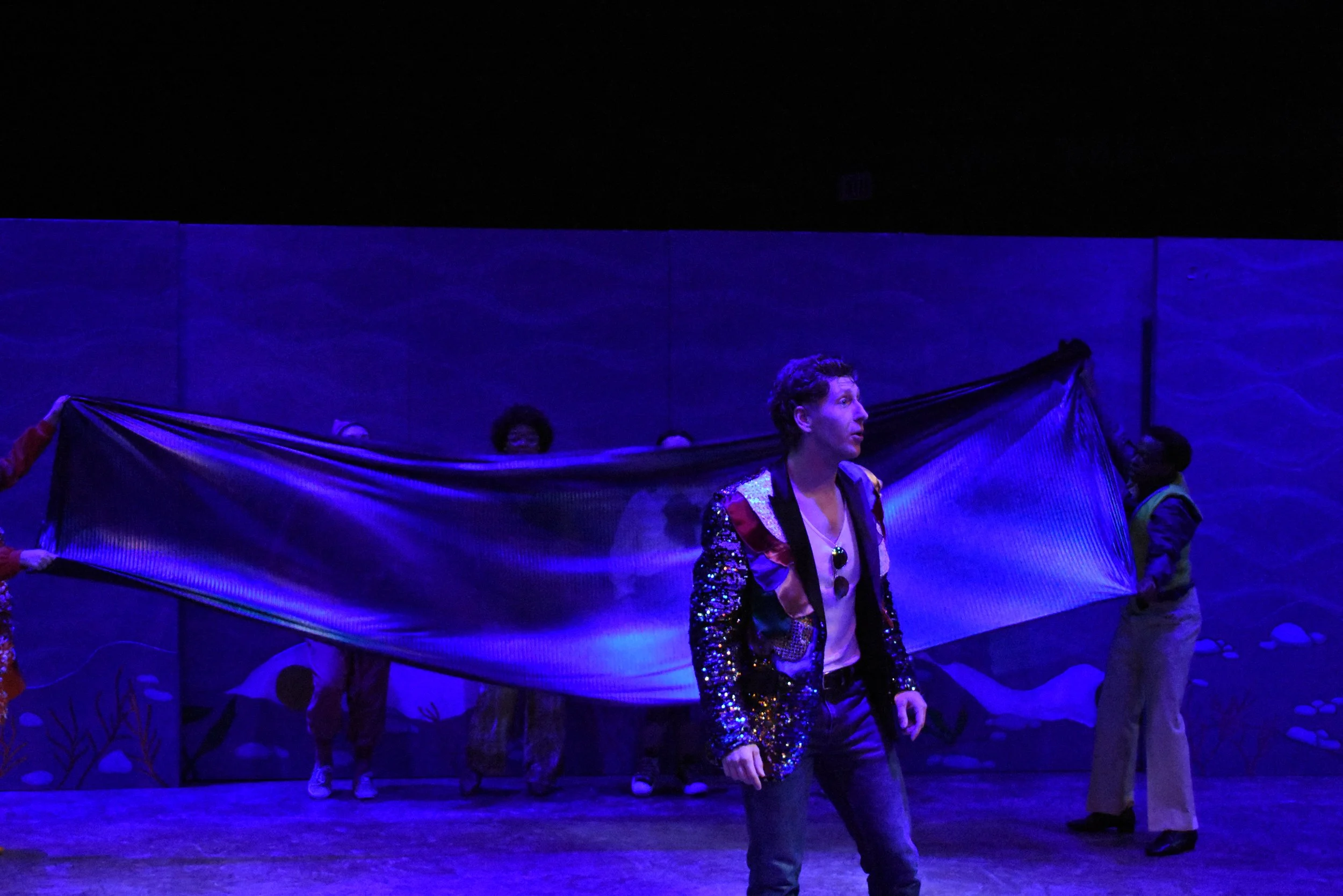 A stage performance with a man in a glittery jacket standing in front of a large blue backdrop, with people holding a long, semi-transparent blue fabric behind him, illuminated with stage lighting.