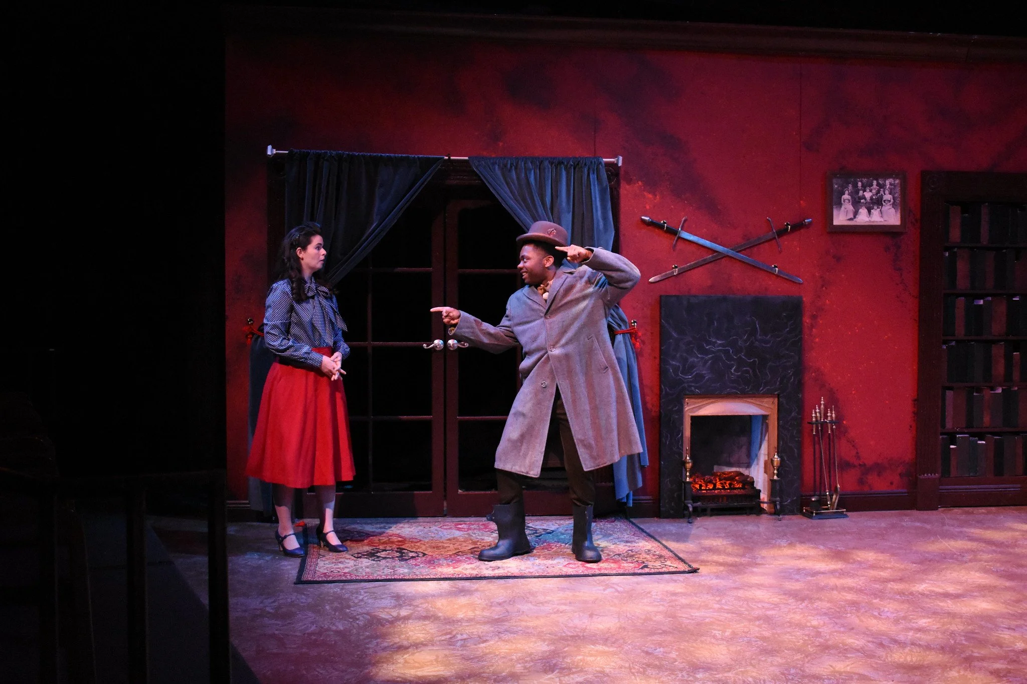 Two actors on stage in a theatrical play: a woman in a blue blouse and red skirt, and a man in a gray coat and hat, standing in a room with a red wall, a fireplace, swords, a picture frame, and bookshelves.