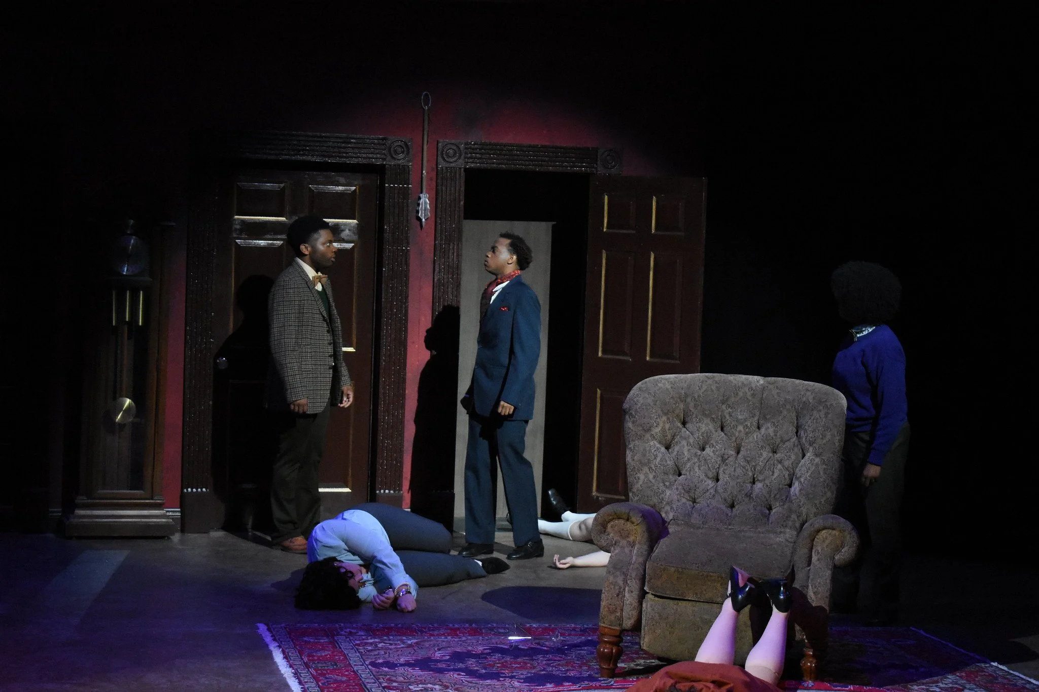 The scene appears to be from a theatrical performance, featuring six characters in a room with dark walls, a vintage armchair, and a red rug. Two men are standing and facing each other near a door, while two women are lying on the floor, one under th