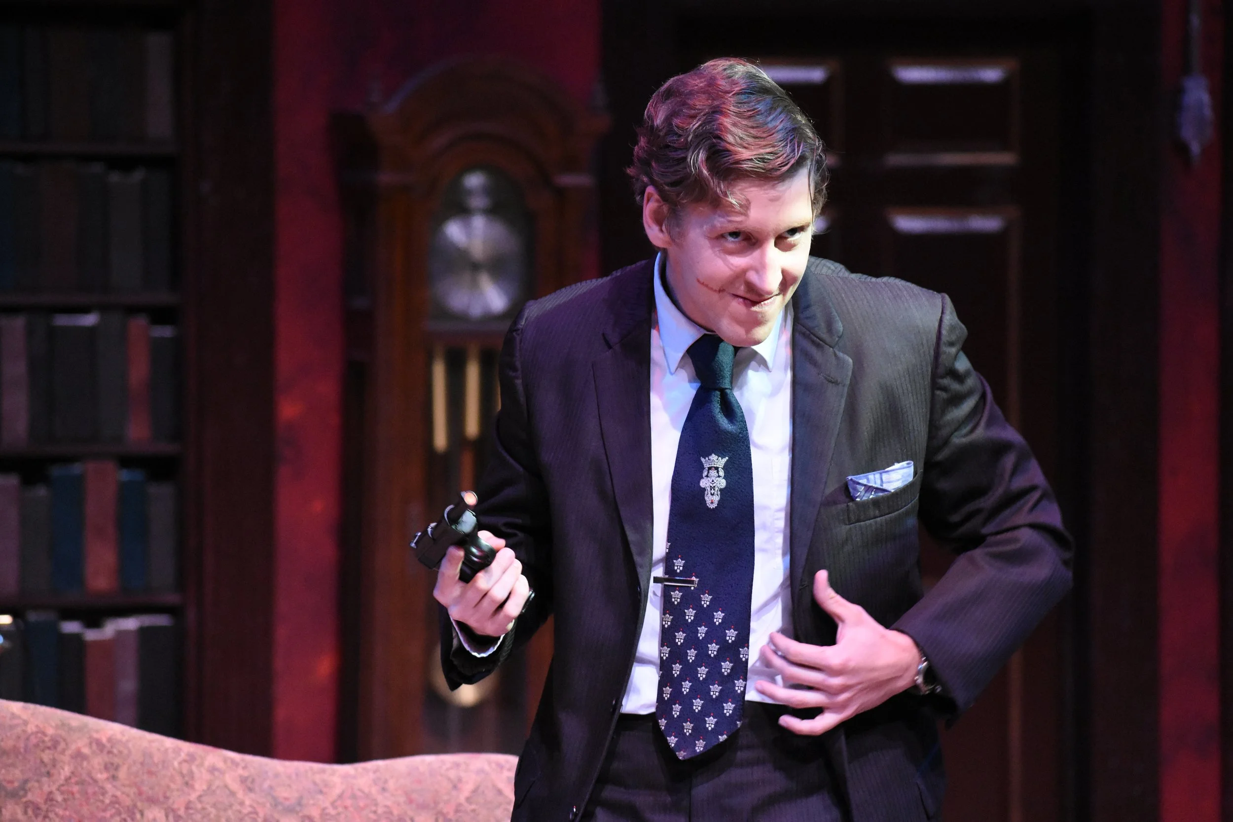 A man in a dark suit holding a revolver in his right hand, with a mischievous smile, in a room with dark wooden bookshelves and a background of a large wooden clock.