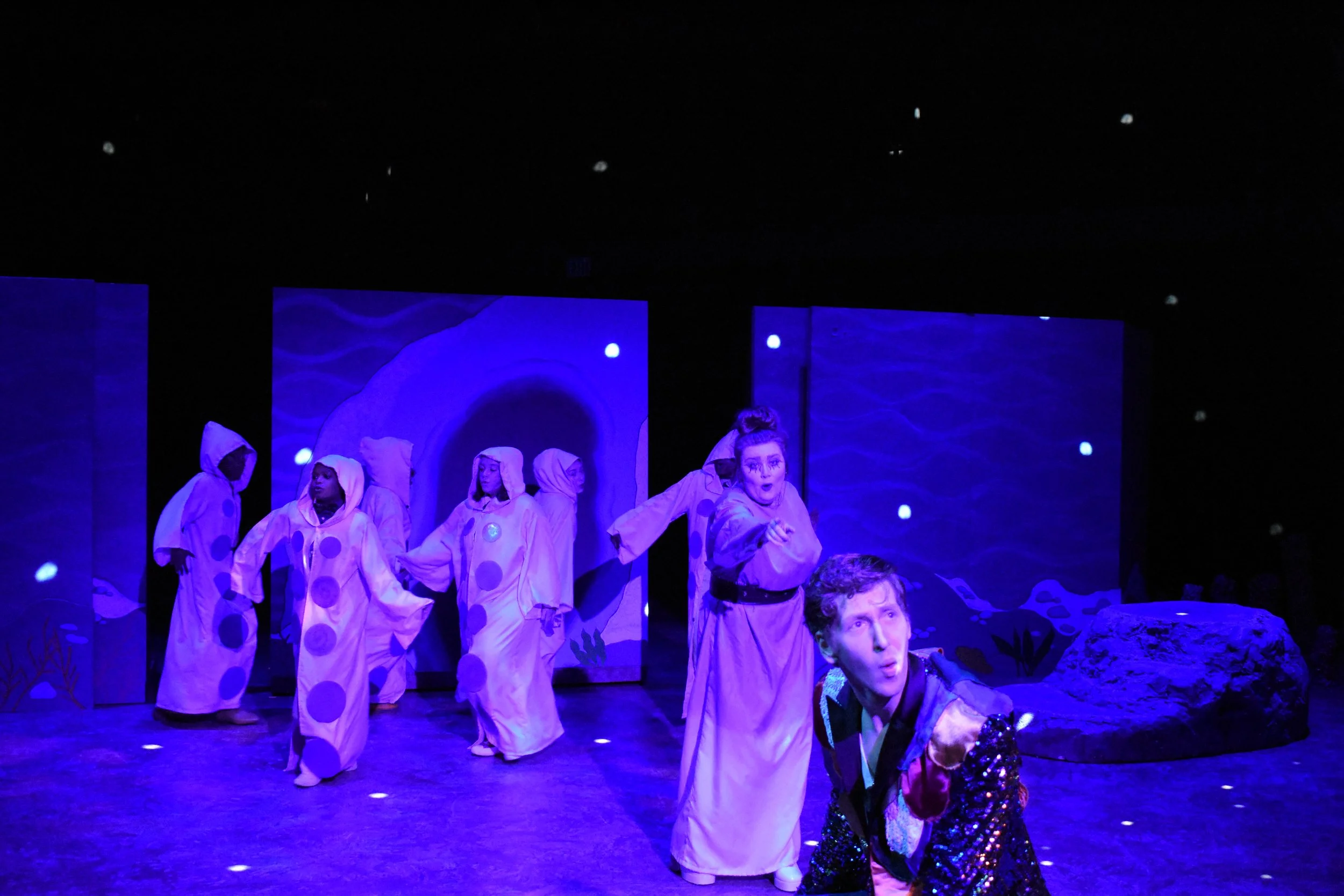 Theatrical performance on stage with seven actors in costumes, with a blue underwater-themed backdrop, under dark lighting.