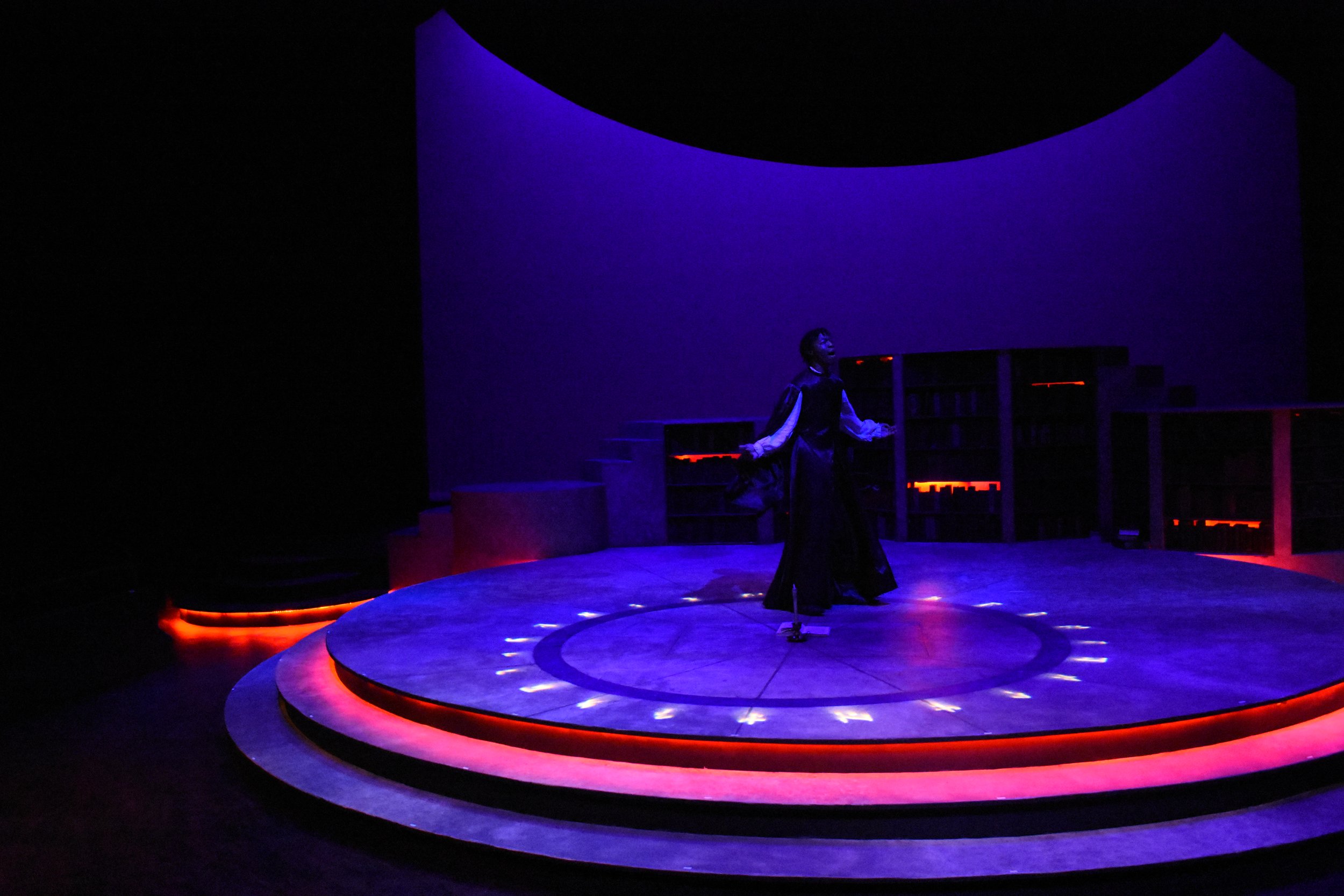 A stage with a woman performing in front of a dark blue and purple background, illuminated by red and purple lighting, with bookshelves in the background.