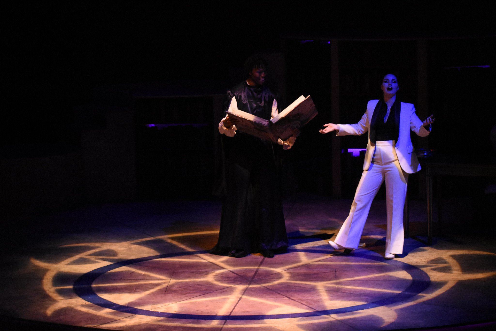 Two performers on a dark stage with a spotlight, one holding a large open book and the other gesturing with hands, with a decorative circular pattern on the floor.