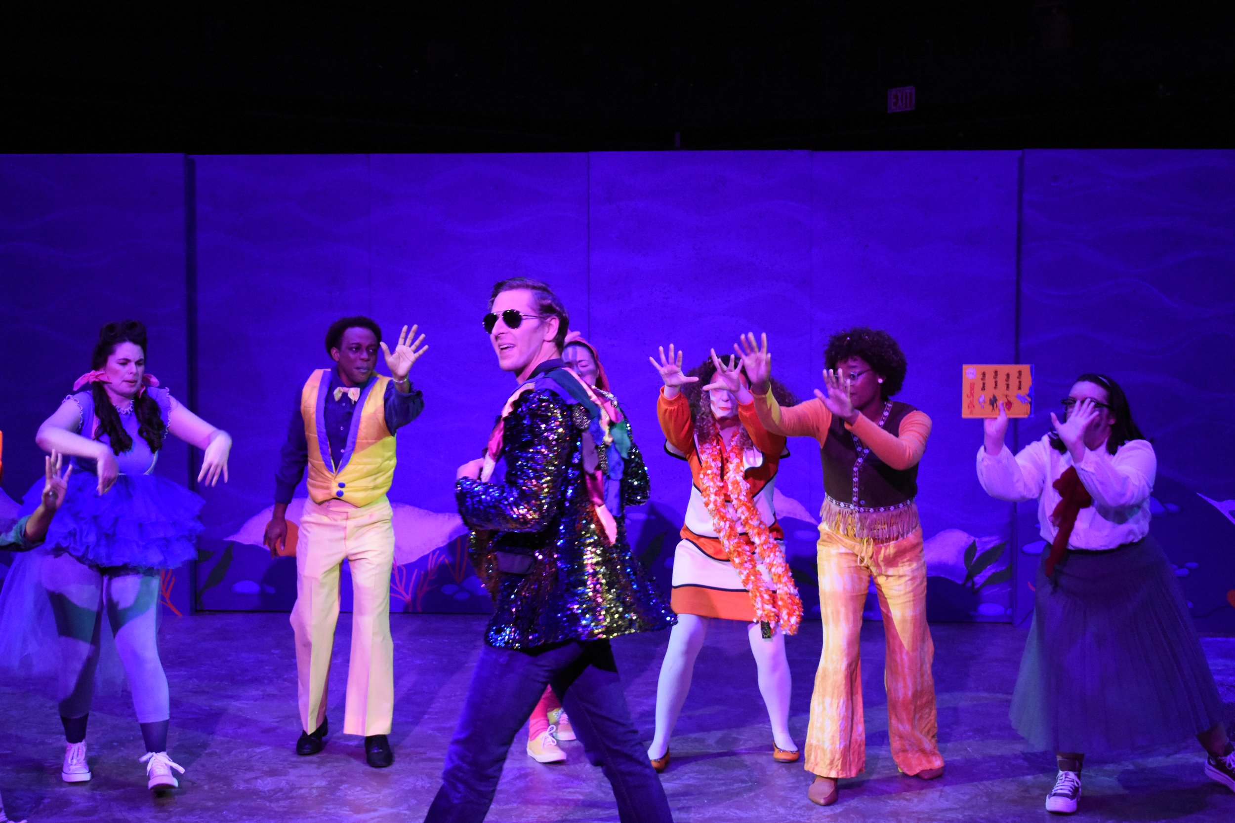 A group of people performing on stage with a purple background, dressed in colorful costumes, some standing and some dancing.