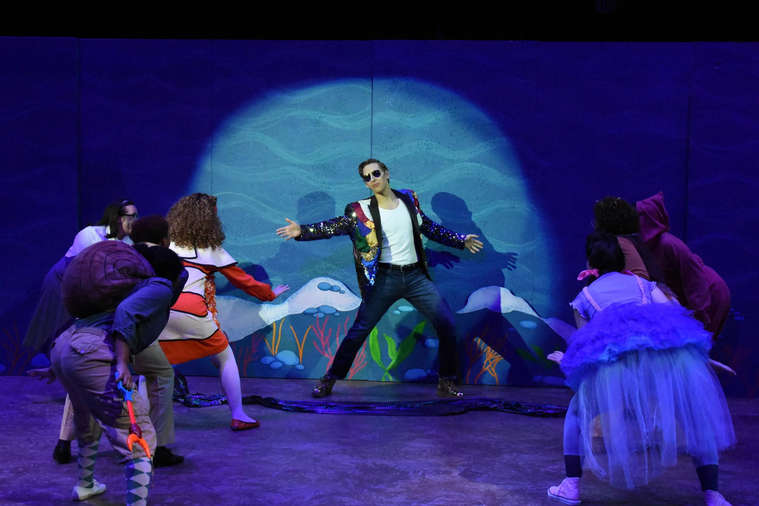 A theater scene with a male performer in a sequined jacket and sunglasses, surrounded by children in costumes, with a colorful underwater backdrop.