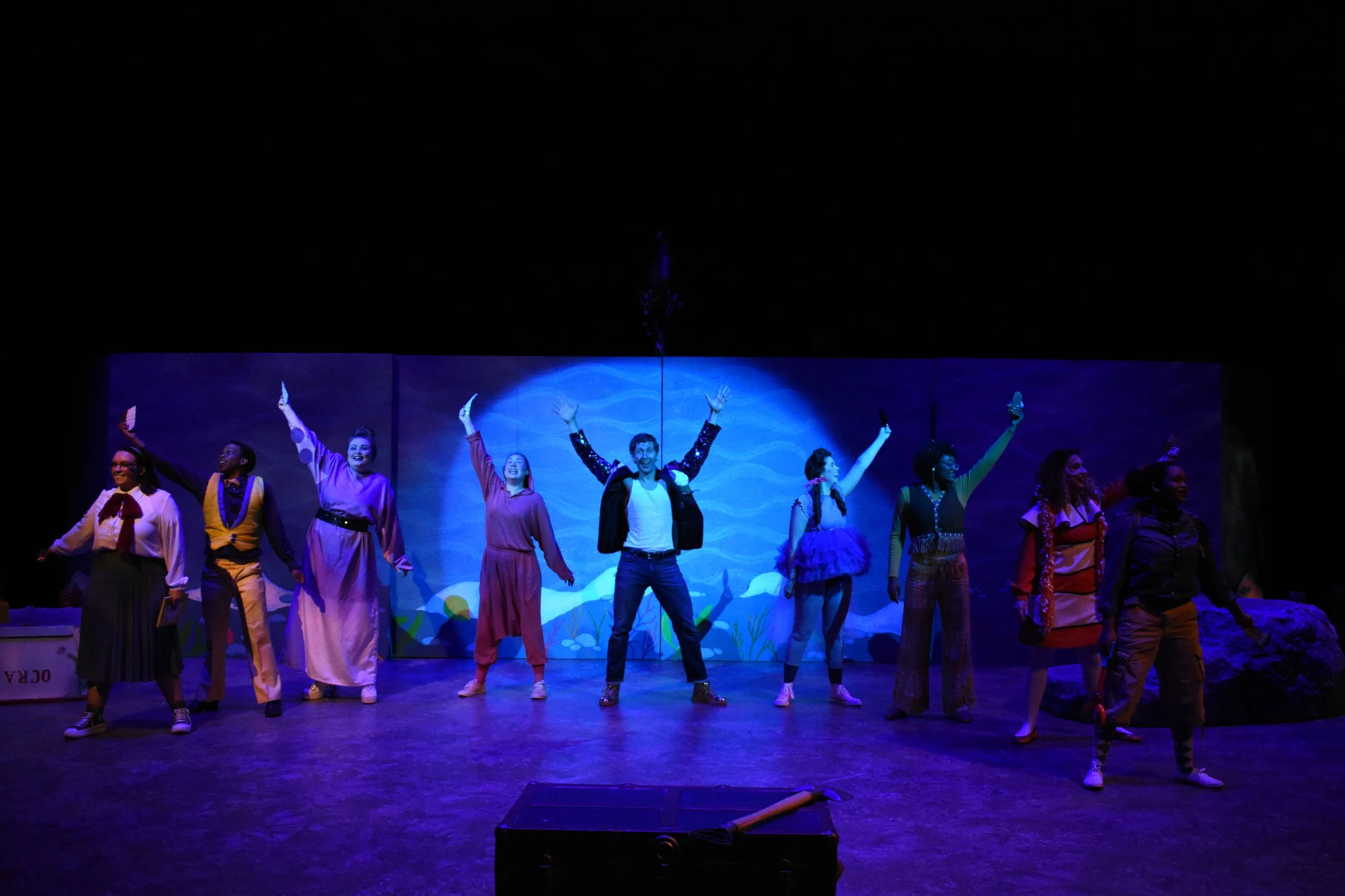 Actors on stage during a theatrical performance, with a blue painted backdrop resembling a lake or ocean scene, and colorful costumes.