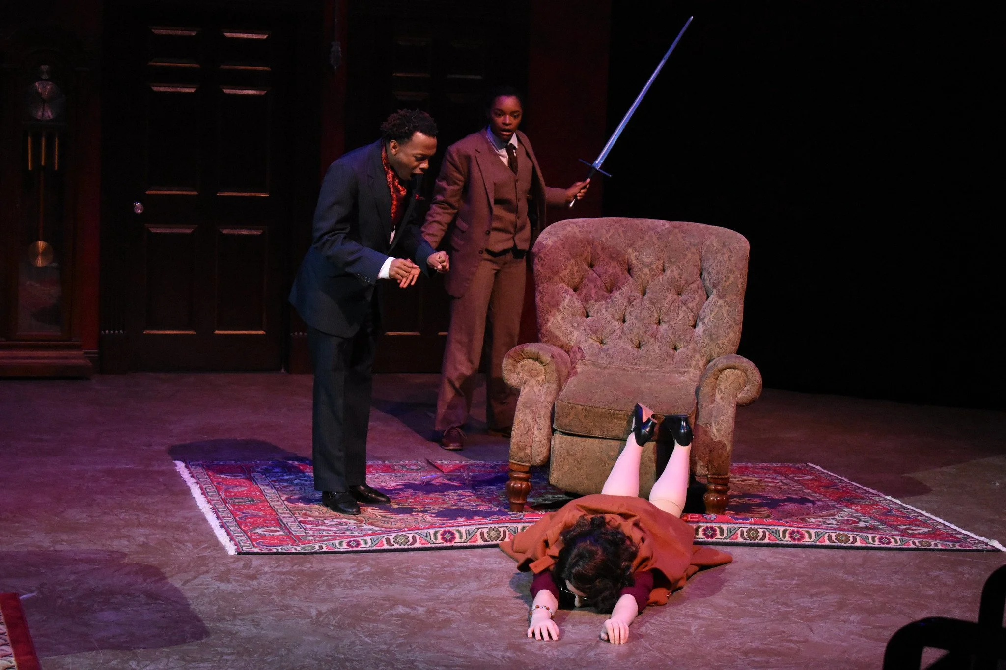 Three actors on stage in a theatrical play, with one lying on the floor and two standing, one holding a sword, in front of a sofa and a wooden cabinet.