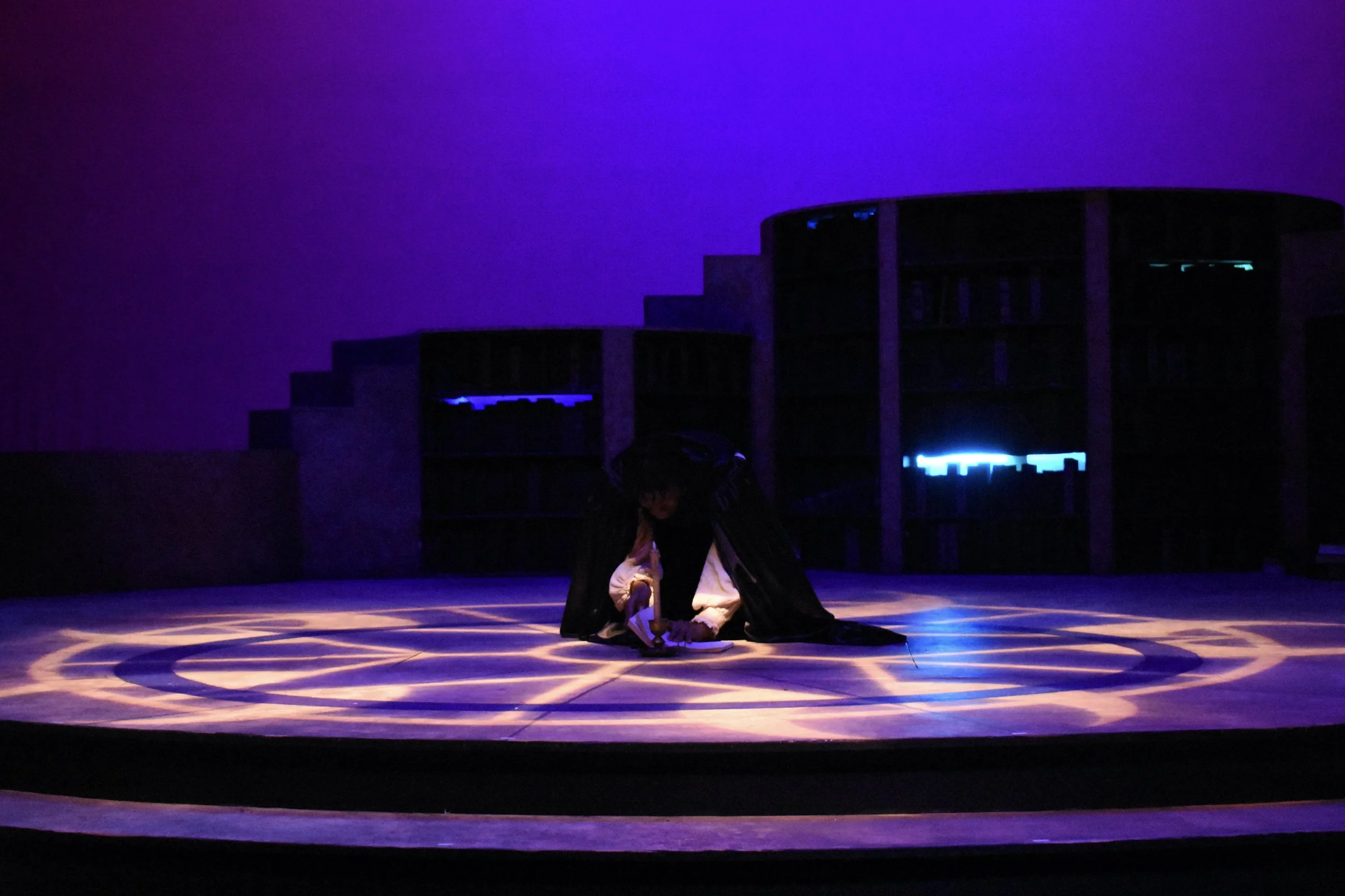 A person dressed as Harry Potter kneeling on a stage with glowing circles, in a dark room with bookshelves and blue lighting in the background.