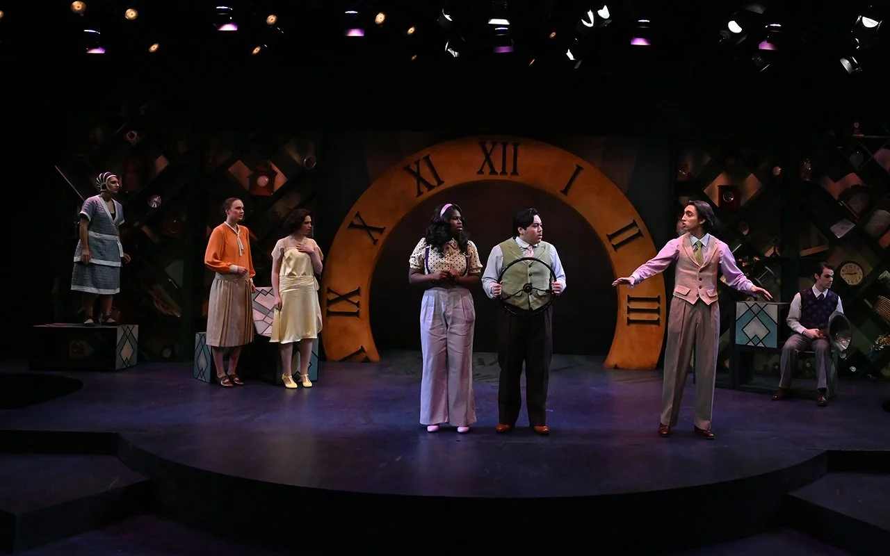 The scene is a theater stage with seven actors performing a play. The background features a large clock face with Roman numerals, and the stage is decorated with bookshelves. The actors are engaged in a theatrical scene, with some standing and others