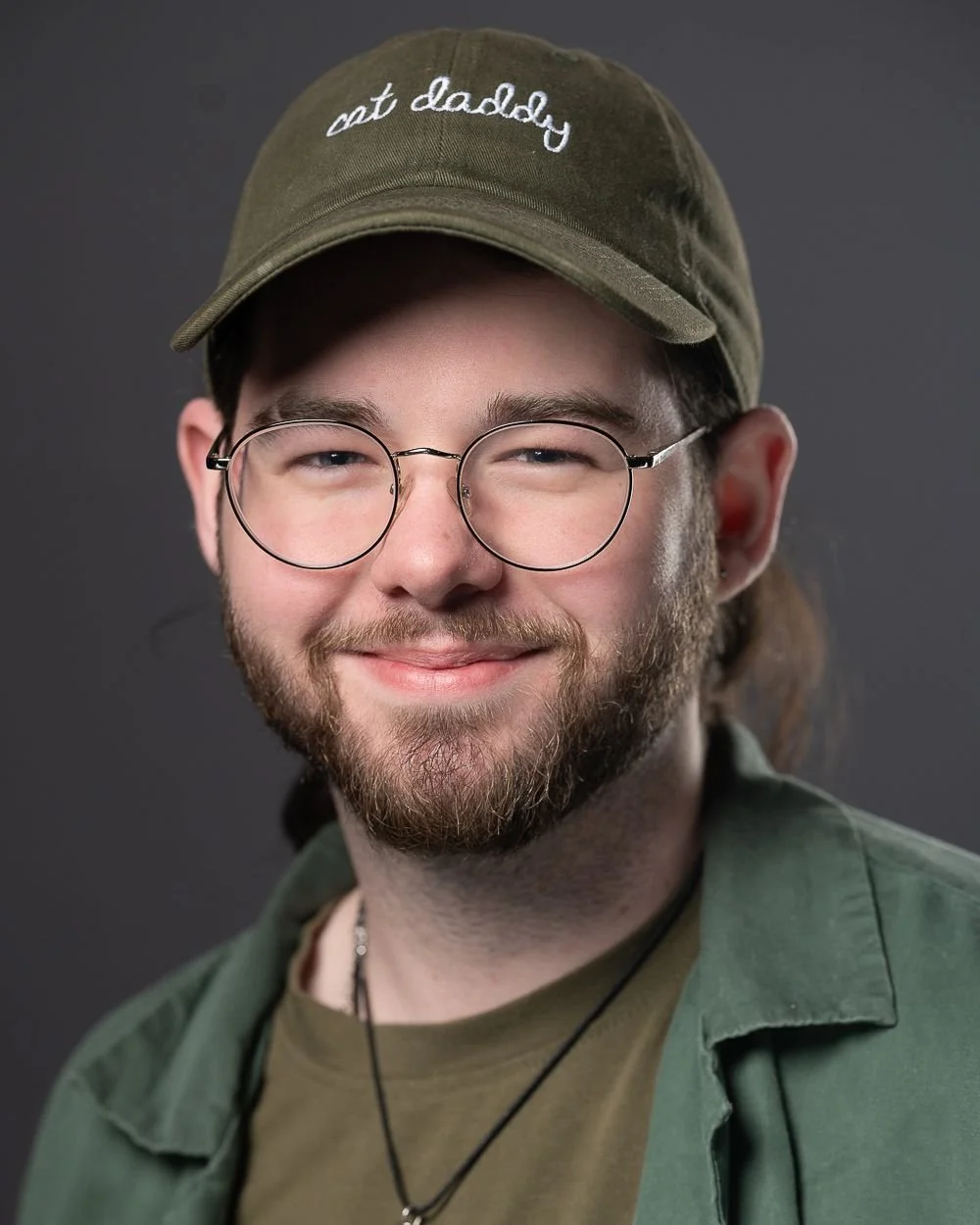 A smiling young man wearing glasses, a green cap with white embroidered text, and casual clothing.