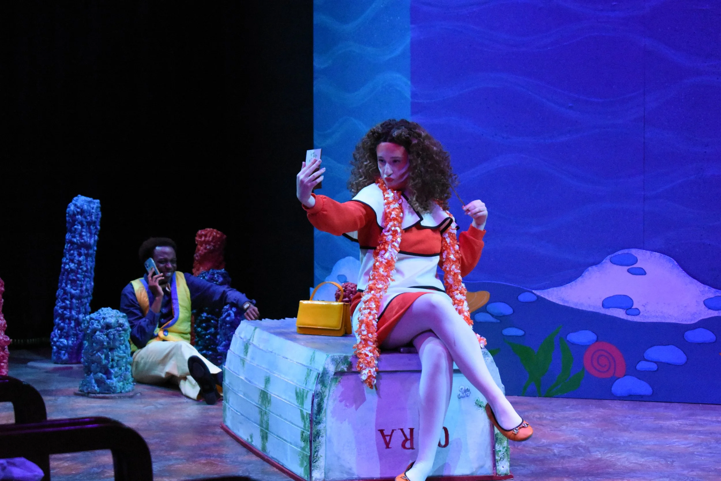 The image depicts a stage scene with a woman with curly hair sitting on a colorful box, wearing a red, white, and black outfit with white stockings and orange shoes. She is holding a phone and taking a selfie. In the background, there are two men, on