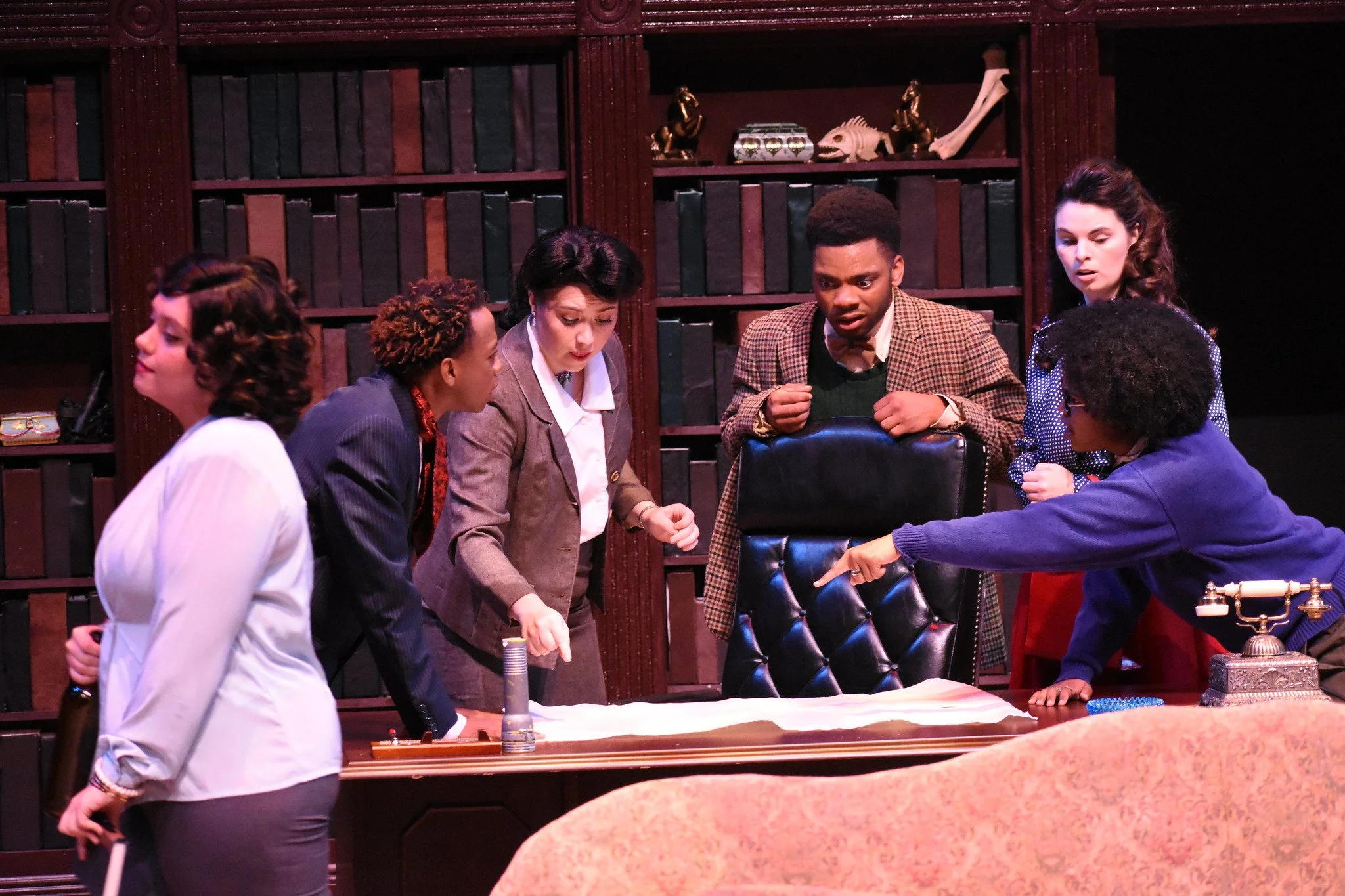 Six actors around a large desk in a library setting, looking surprised or intense, with shelves of books and decorative items behind them.