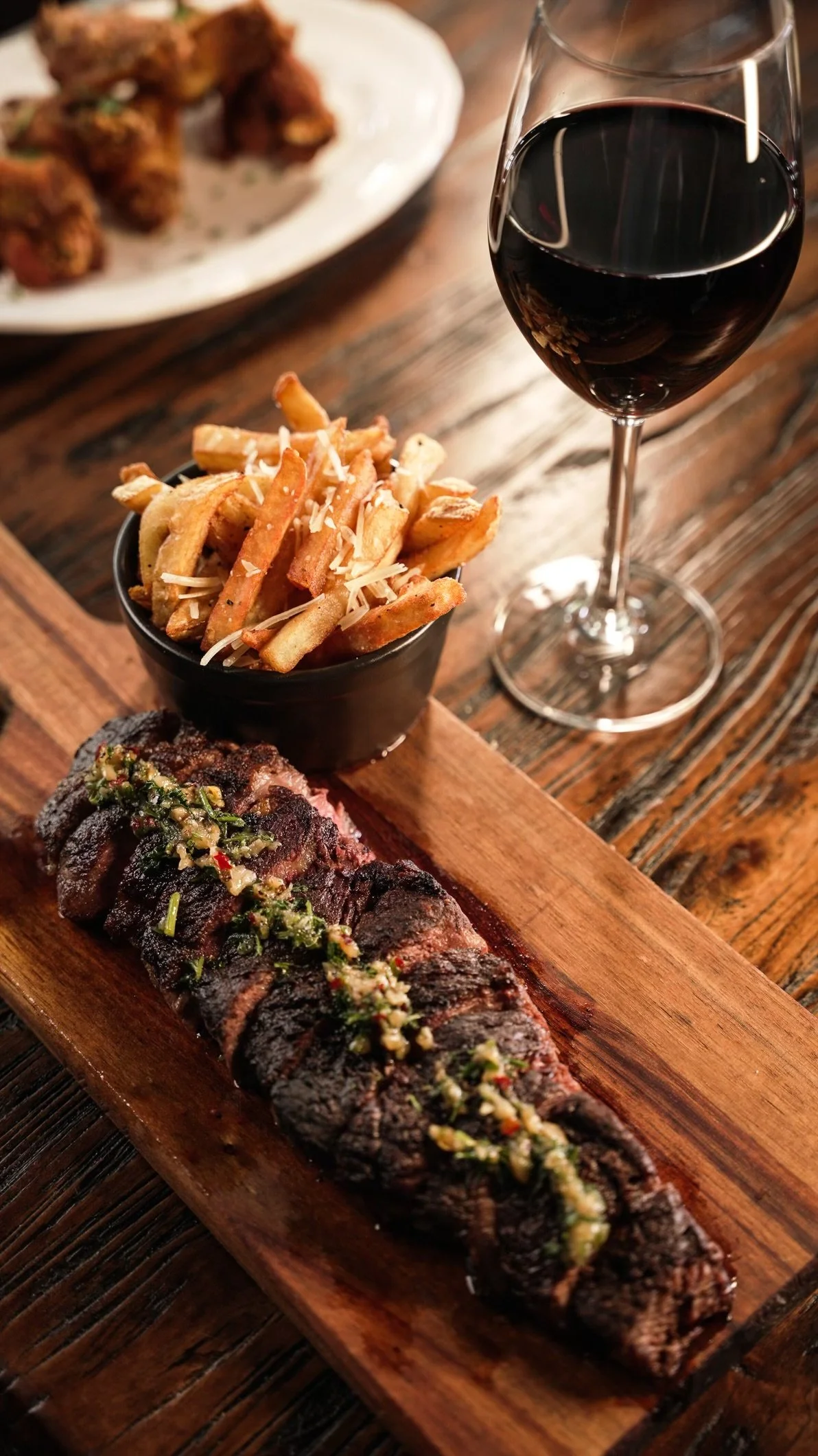 Close-up of a wooden serving board with a grilled steak topped with chopped herbs and garlic, a small bowl of French fries with cheese, and a glass of red wine, with a dish of fried chicken in the background.