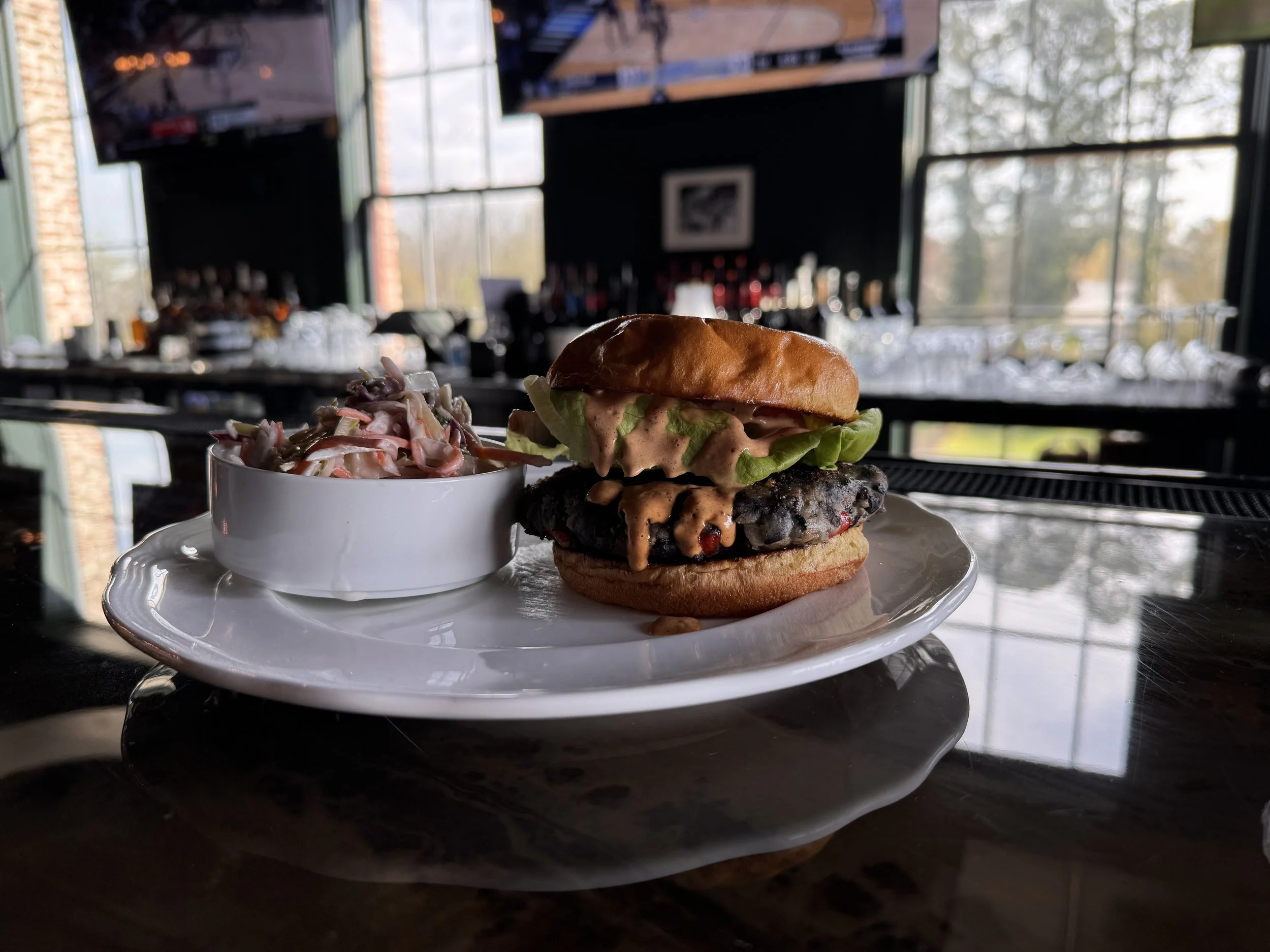 A burger with lettuce, sauce, and a beef patty on a bun, served with a side of coleslaw on a white plate at a bar or restaurant.