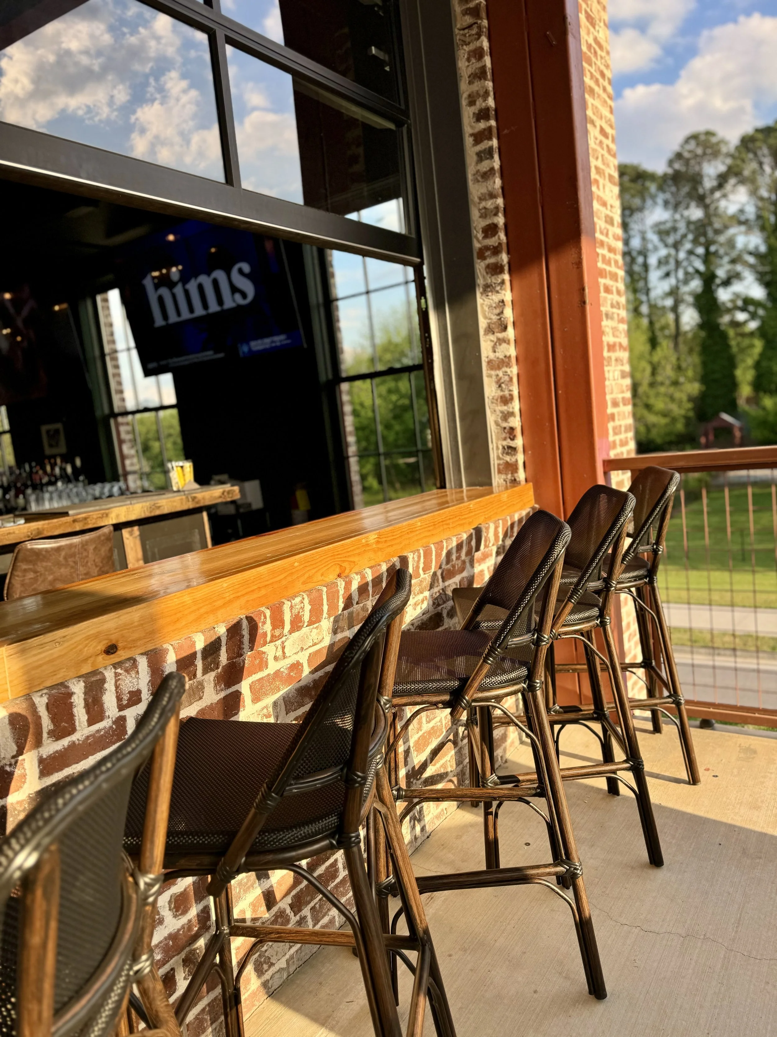 Sunlit outdoor bar area with wooden countertop and black metal bar stools in front of a brick wall, overlooking a green landscape and blue sky with clouds.