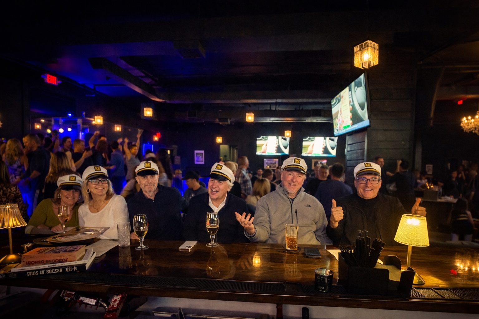 A group of six older adults wearing captain hats sitting at a bar counter, smiling, with drinks in front of them in a lively bar or restaurant with many people.