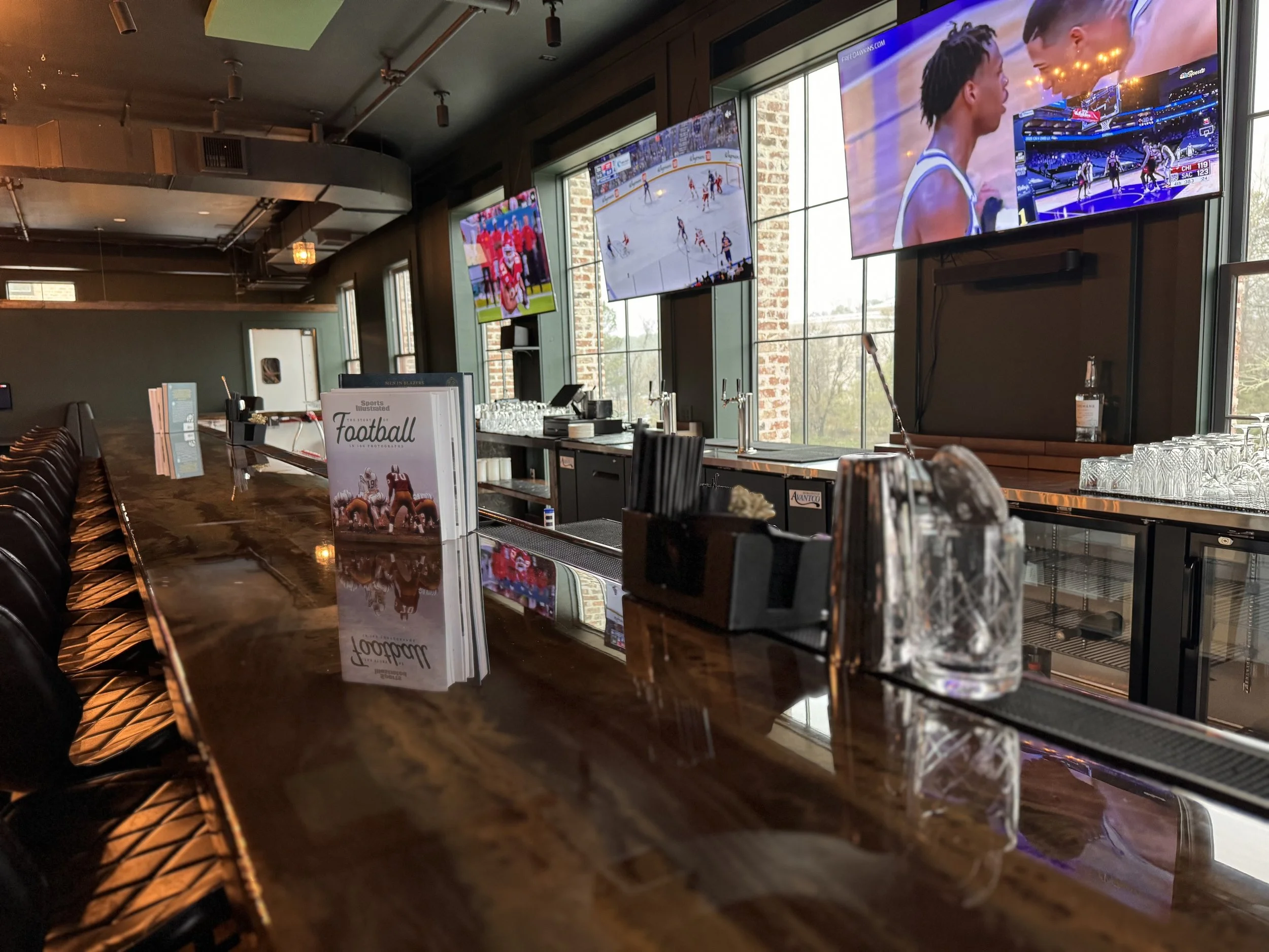 Empty bar with a dark wooden counter, menu on display, and multiple television screens showing sports games inside a sports bar or restaurant.