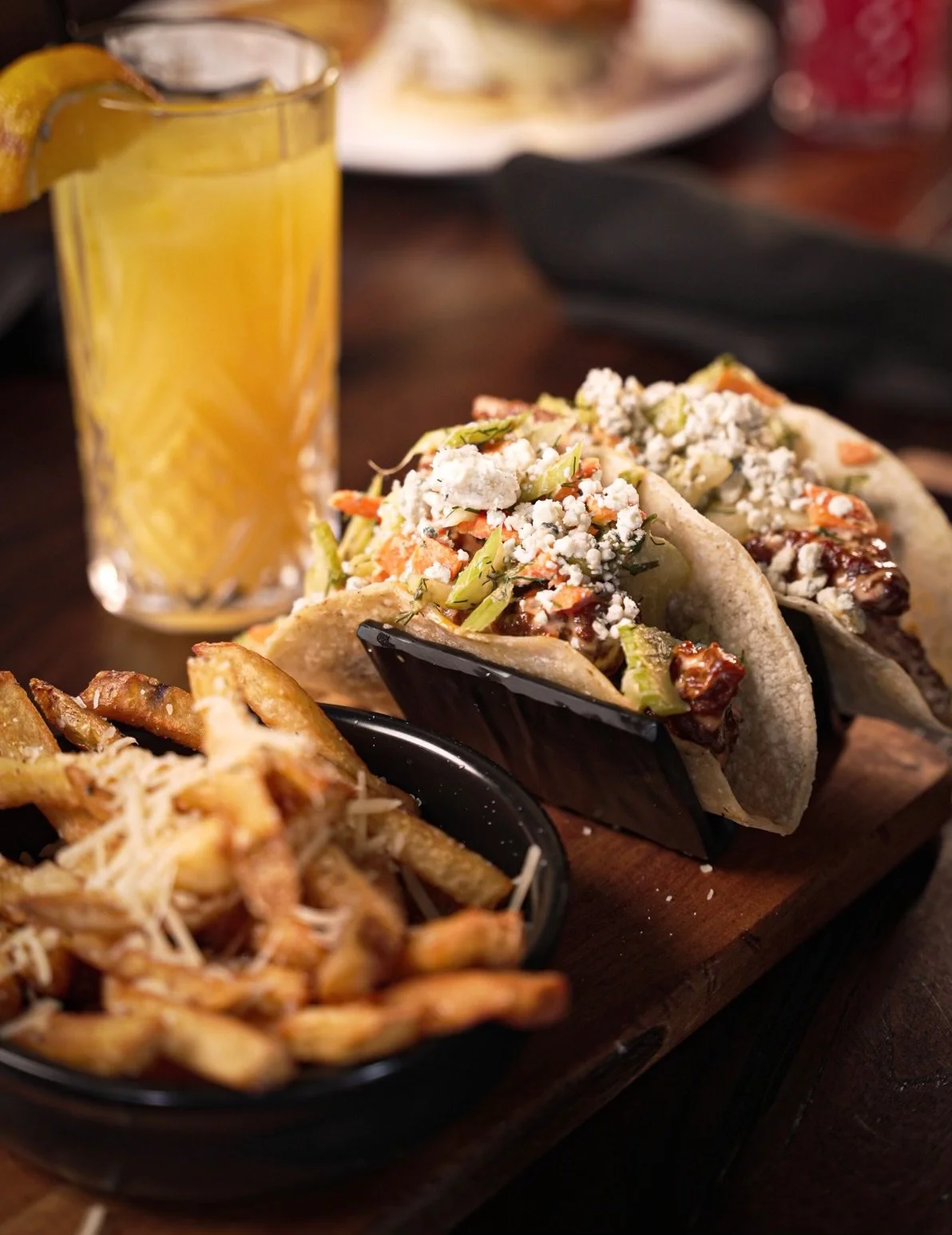 Three tacos filled with shredded lettuce, cheese, vegetables, and sauce, served on a wooden board with a side of French fries and a glass of orange juice.
