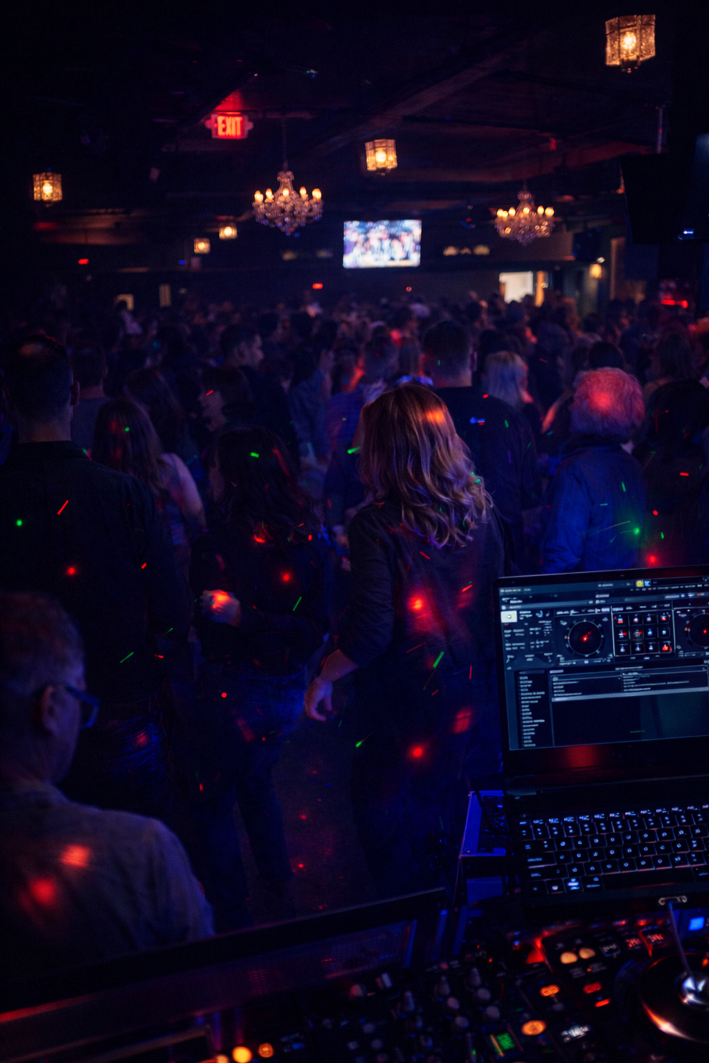 A crowded nightclub dance floor with colorful lights, chandeliers, and a DJ booth with a laptop in the foreground.