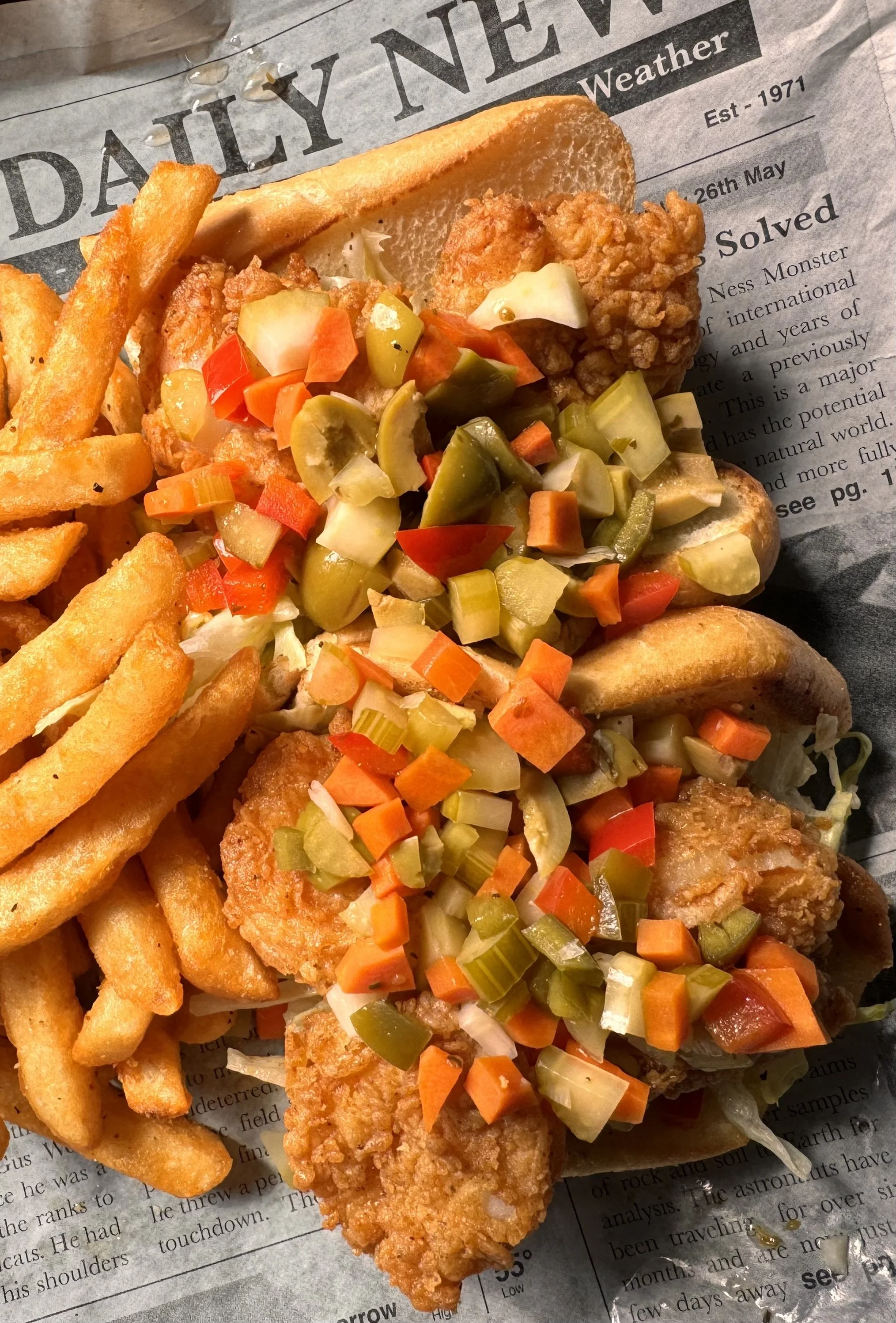 Fried fish sandwich with pickles, coleslaw, French fries, and a side of potato wedges on newspaper