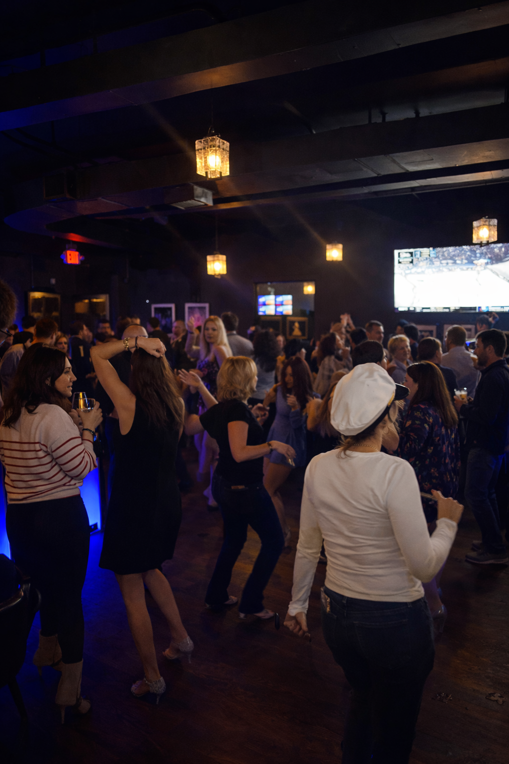 People dancing and socializing in a dimly lit bar or nightclub with hanging pendant lights and large screens on the wall.