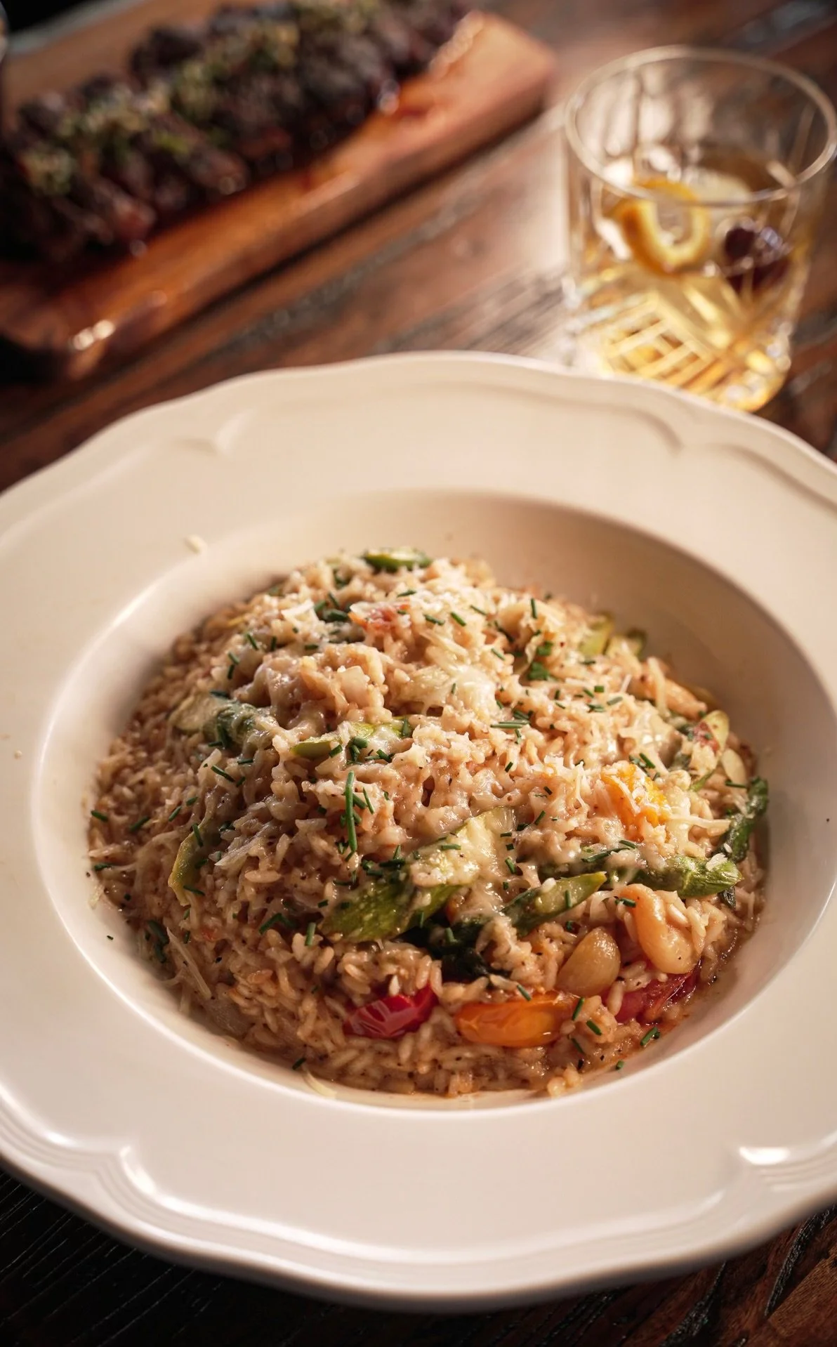 A bowl of cooked risotto with vegetables, garnished with chopped herbs, on a white plate. In the background, a glass of white wine with lemon slices and a wooden board with dark chocolates.