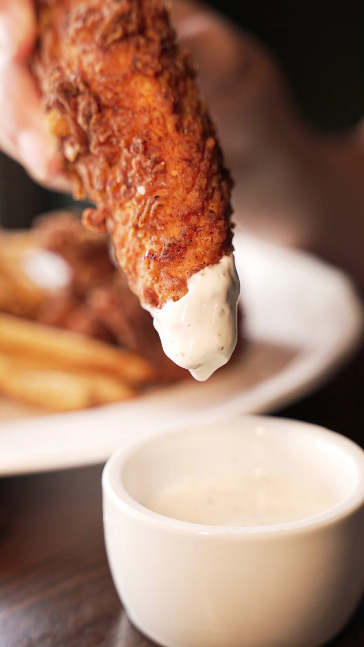 Close-up of a fried chicken drumstick being dipped into white creamy sauce, with a plate of fries in the background.
