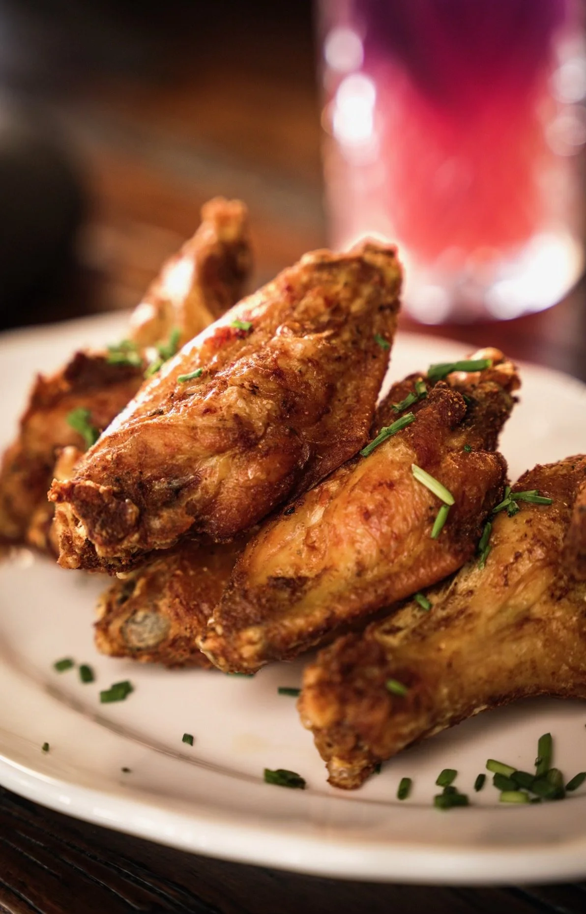Close-up of crispy fried chicken drumsticks garnished with chopped herbs, placed on a white plate with a glass of red drink in the background.