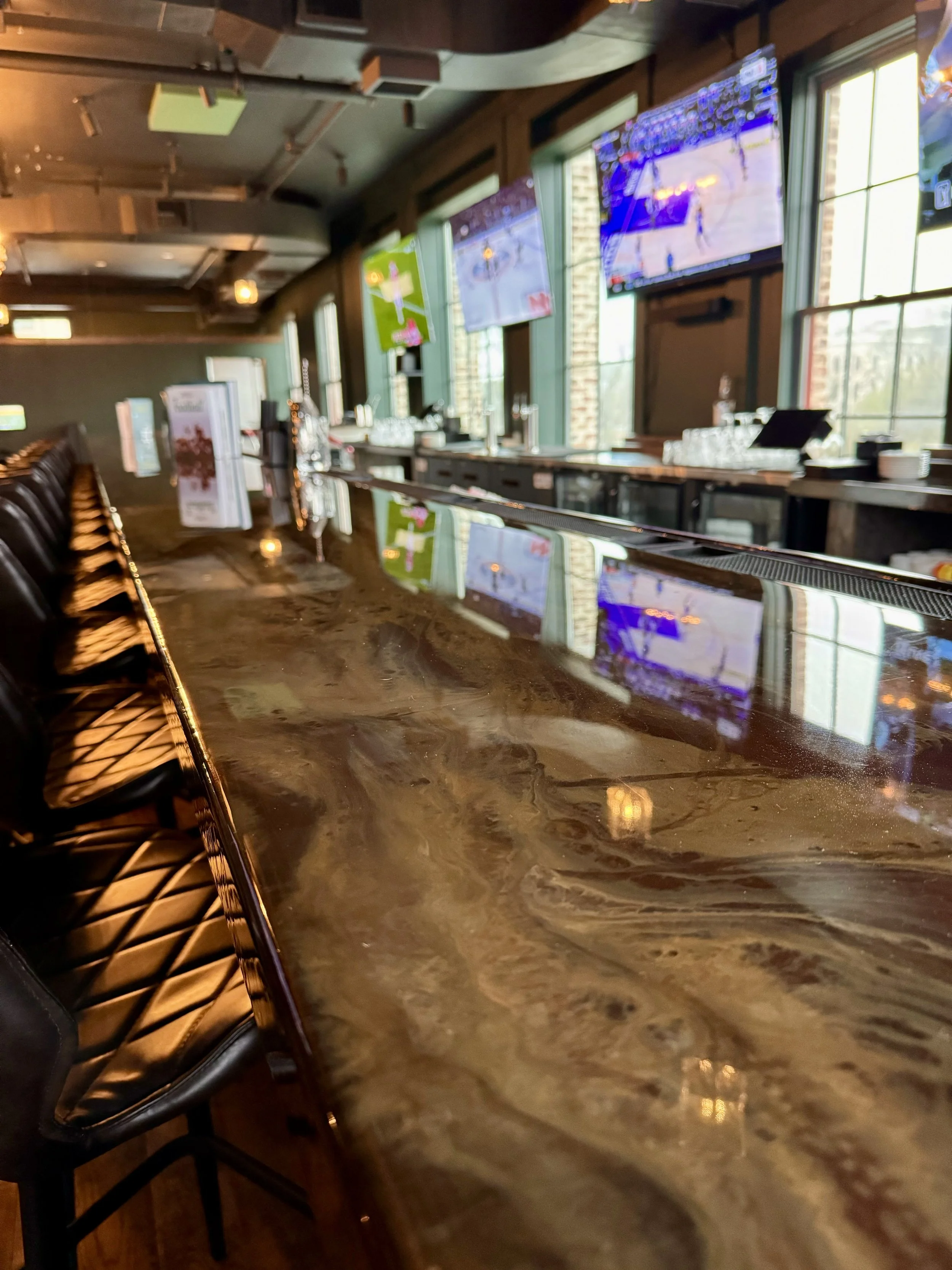 Empty bar with a polished marble countertop and black leather bar stools in a sports bar with multiple large TVs showing sports games, and large windows letting in natural light.