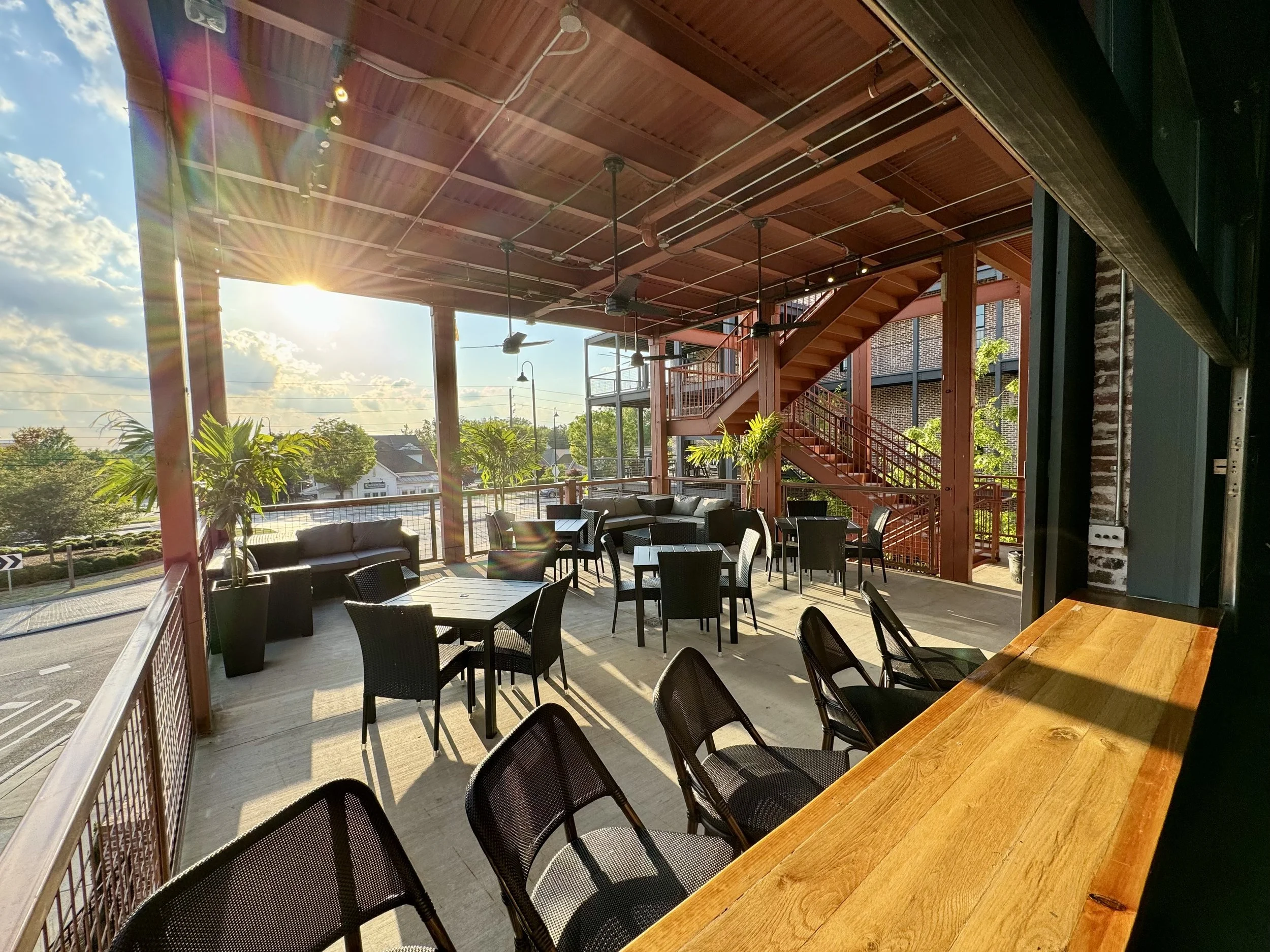 Empty outdoor patio with tables, chairs, potted plants, staircase, and sunlight streaming in.
