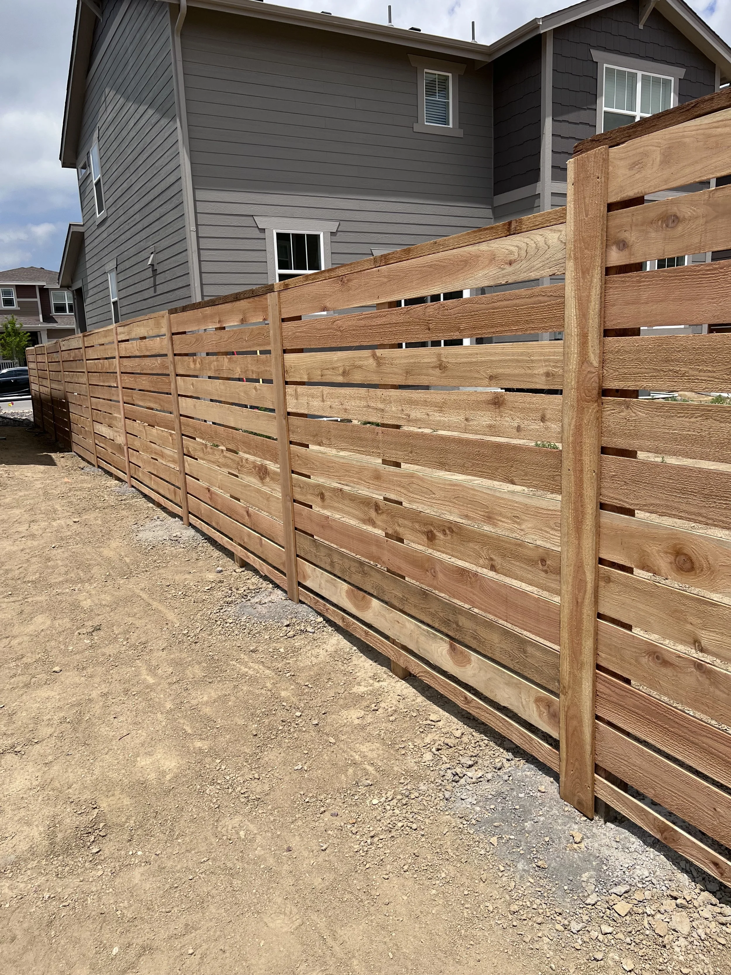New wooden privacy fence along a dirt yard in front of a gray two-story house with multiple windows.