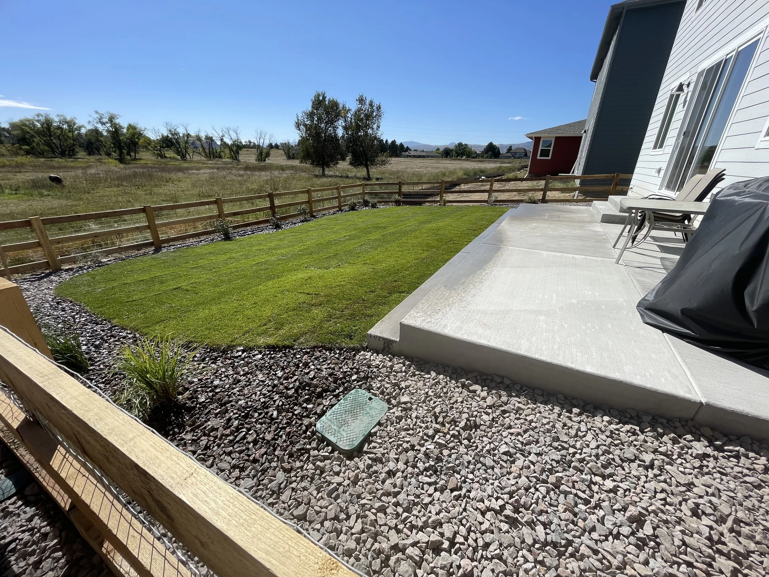 Backyard with a newly laid green lawn, concrete patio, white house siding, outdoor chairs, charcoal grill covered with black cover, gravel border, wooden fence, open field and trees in the background under blue sky.