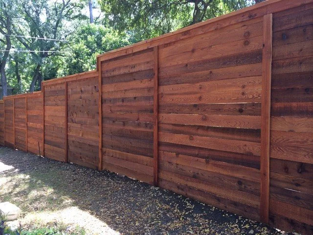 A newly constructed wooden privacy fence with horizontal planks, installed in a backyard with trees in the background.
