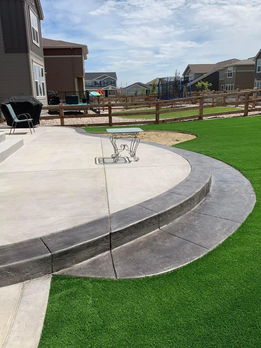 Residential backyard with a concrete patio, artificial grass or turf, a small staircase, a black barbecue grill, a chair, a table, a fenced yard, and a swing set in the background under a partly cloudy sky.