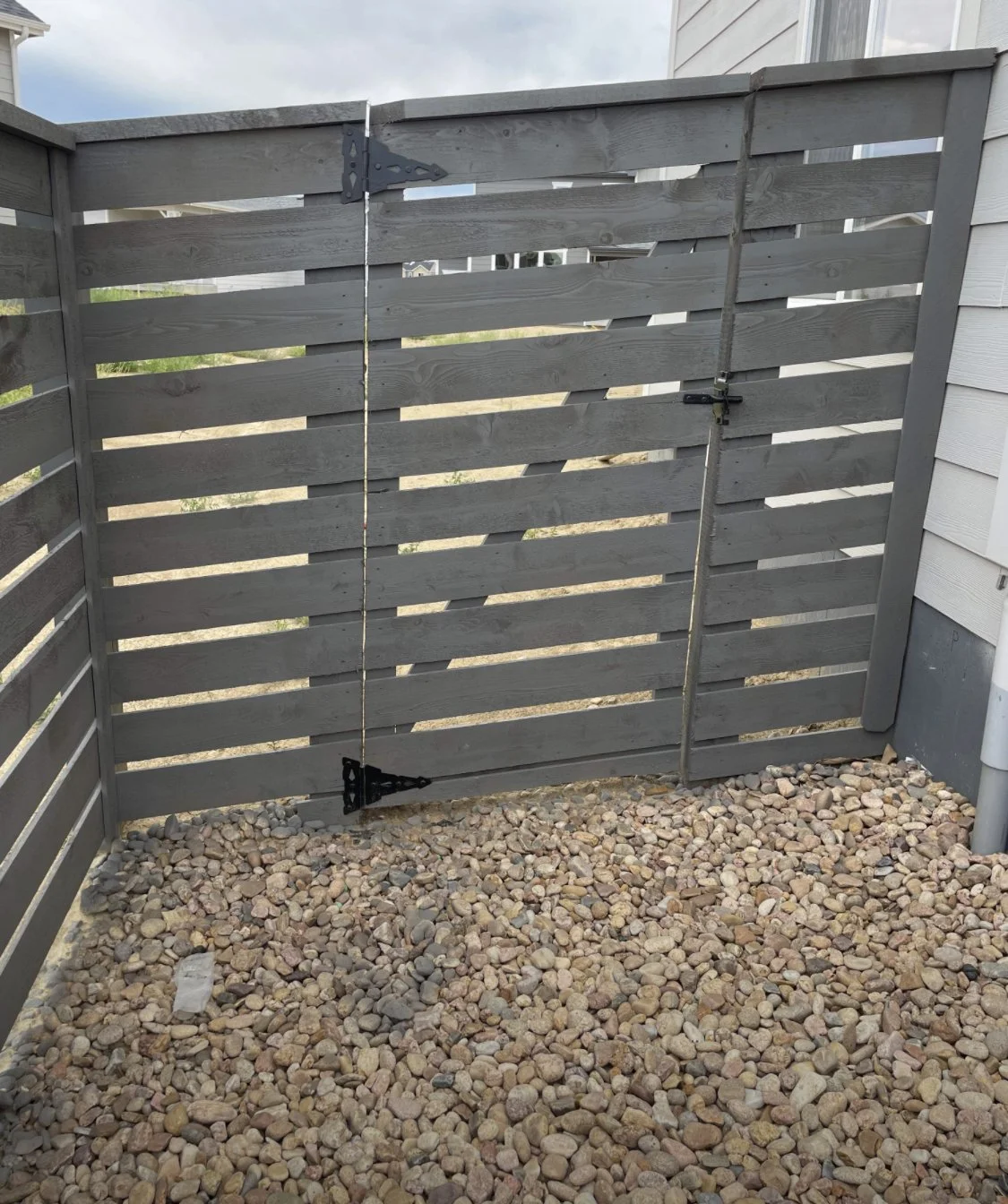 Gray wooden privacy gate with a diagonal latch and a vertical rope tensioner on a gravel ground, attached to a gray wooden fence on both sides.