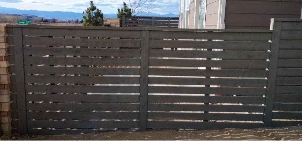 A wooden privacy fence with horizontal slats in front of a backyard, with a house wall on the right and trees and mountains in the distance.