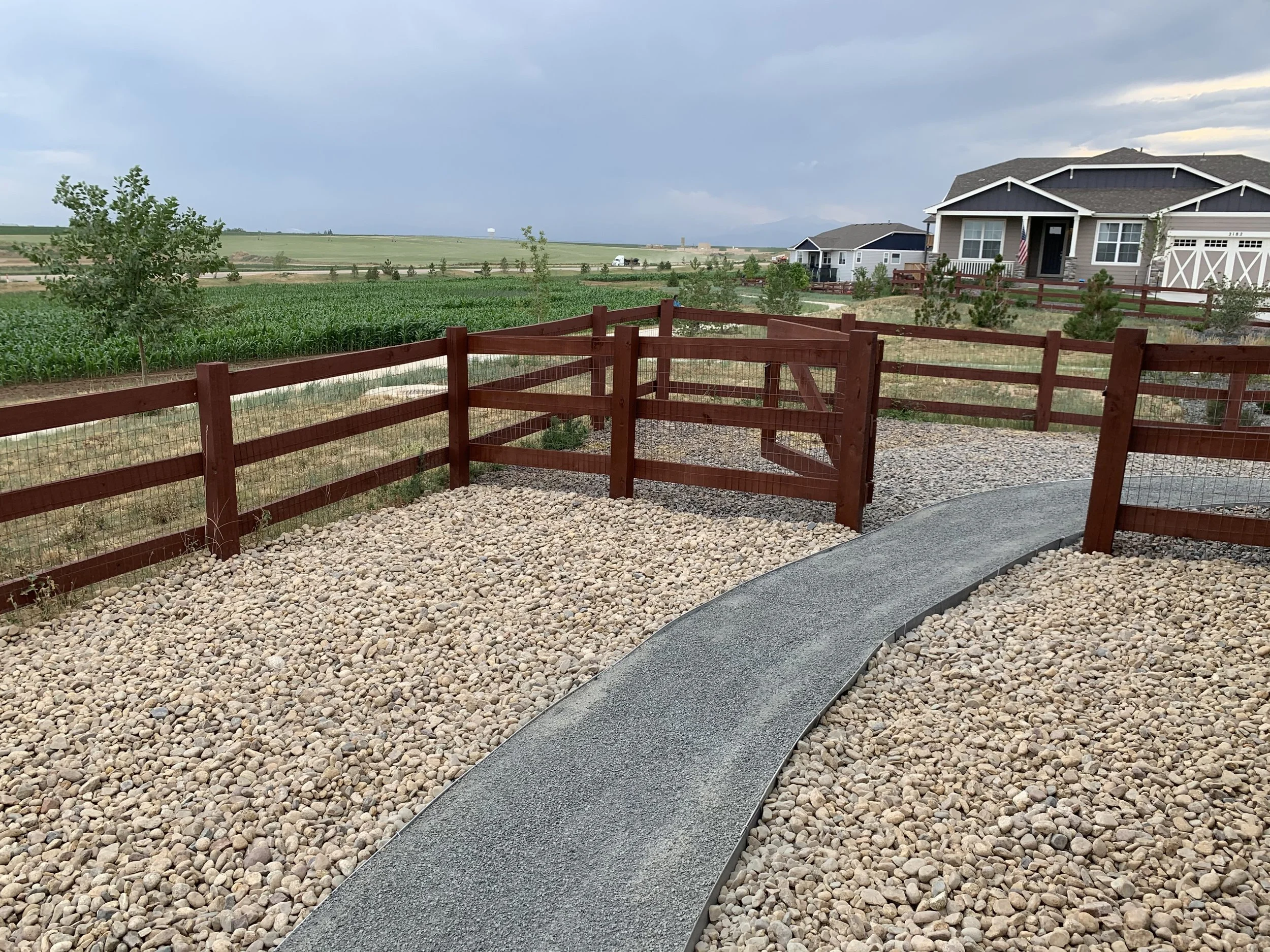 Residential backyard with a winding gravel pathway, wooden fence, and suburban houses in the background under a cloudy sky.