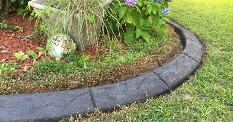 A garden bed with plants, mulch, and decorative ceramic objects, bordered by a black colored stamped concrete curbing separating the mulch bed from a grassy lawn.
