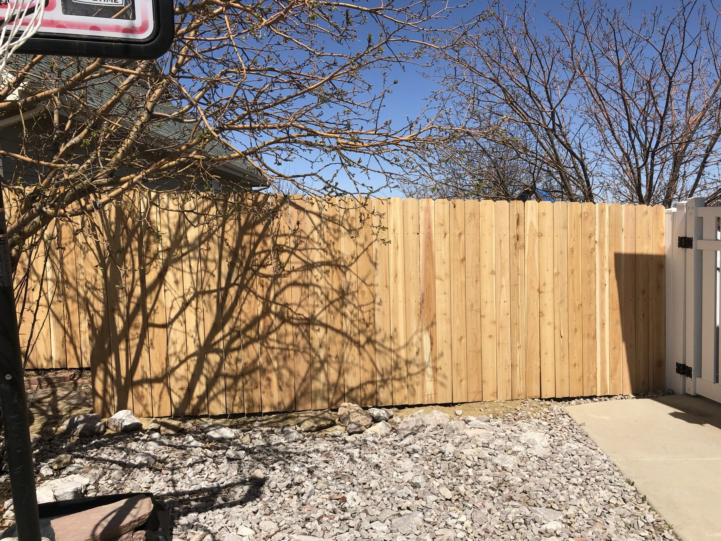 A wooden privacy fence surrounds a backyard. There is a tree with no leaves casting shadows on the fence. The ground has gravel and a concrete walkway. Part of a white gate is visible on the right side. A house roof and a partly cloudy blue sky are i